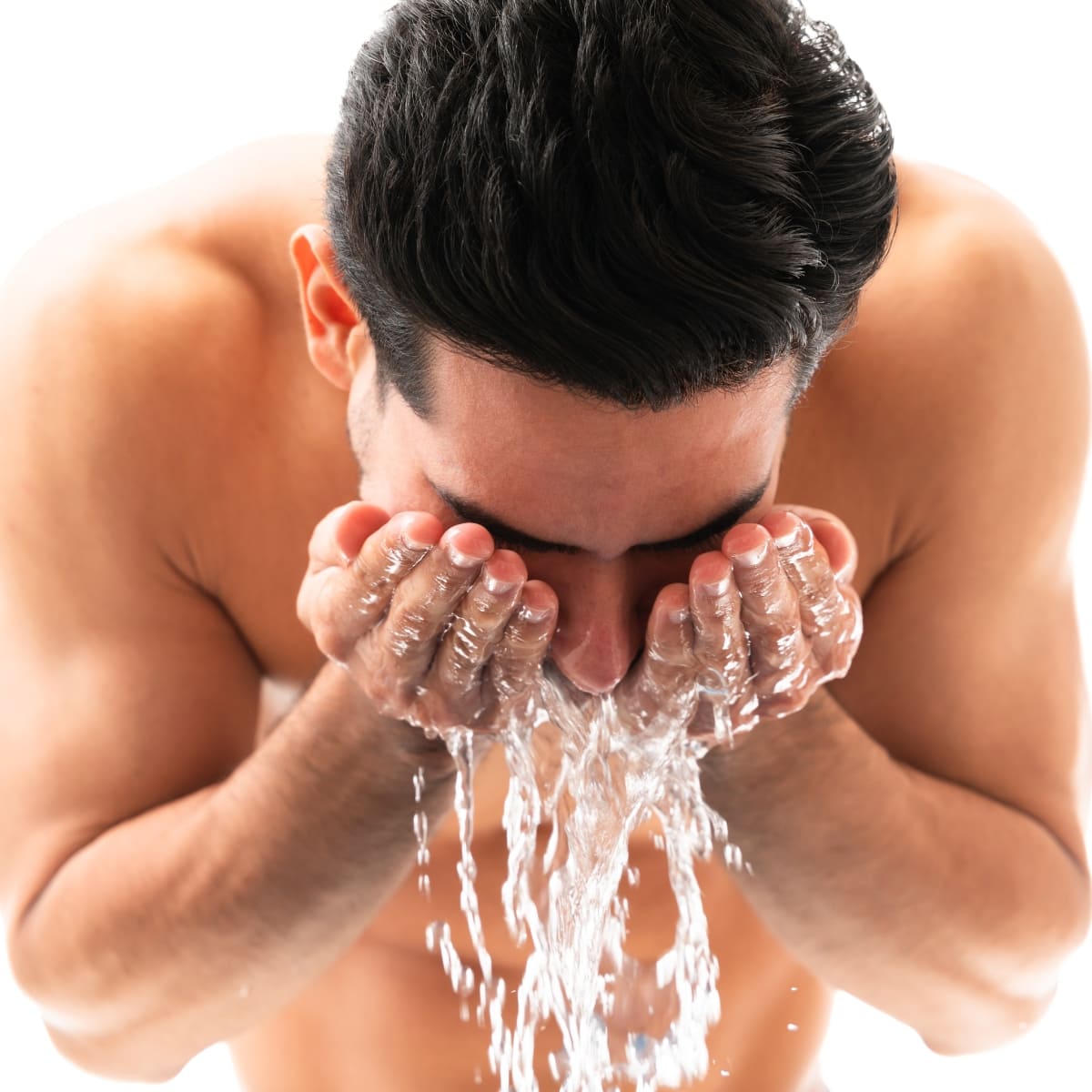 A shirtless man with dark hair is bent over while washing his face. He is cupping water in his hands and splashing it onto his face, with water visibly dripping down. The background is a plain, light color, emphasizing the refreshing act of face washing as part of his grooming routine.