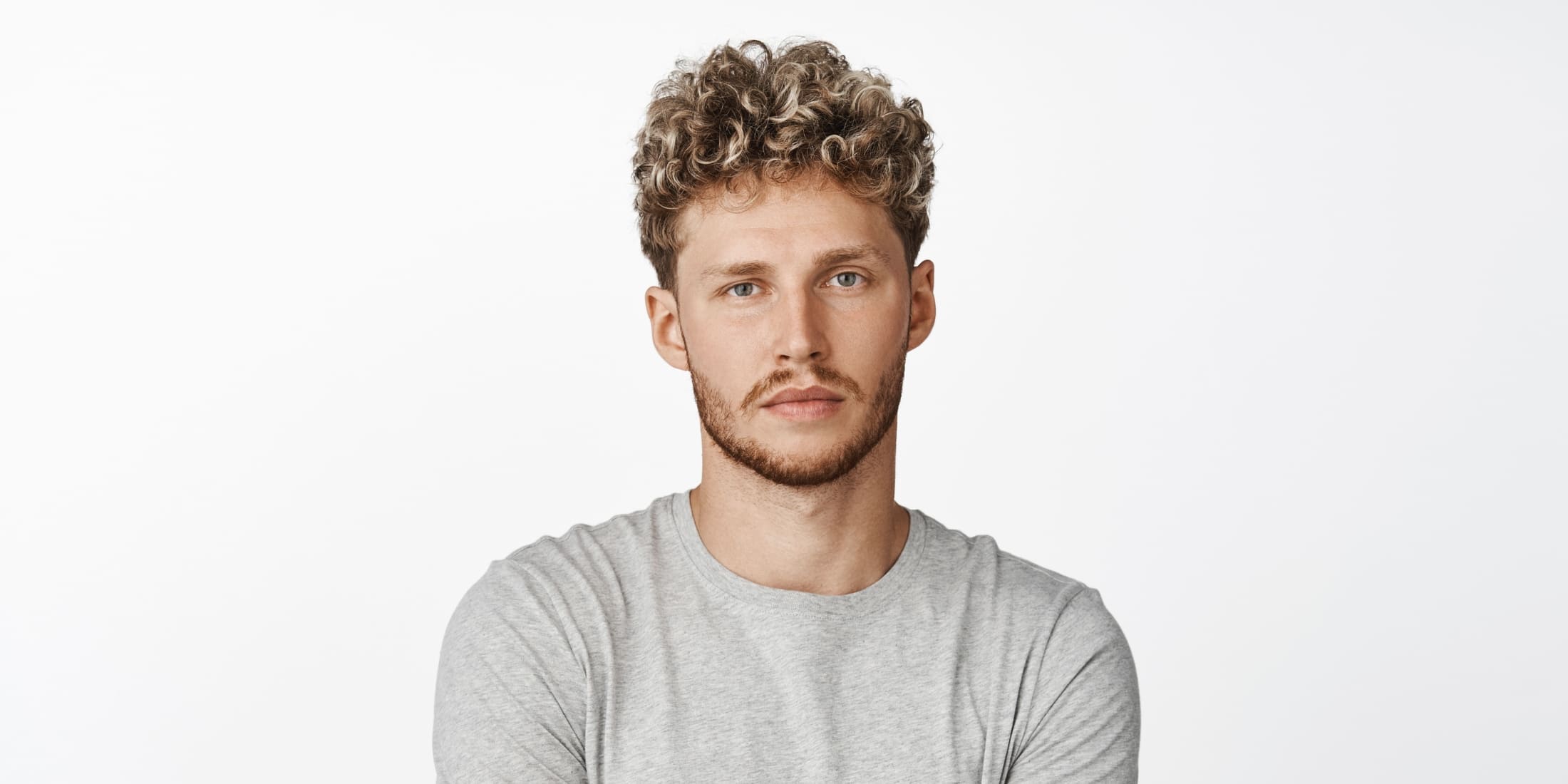 A young man with curly hair and a light brown, 2 week beard is looking directly at the camera with a neutral expression. He is wearing a light gray t-shirt and stands against a plain white background. His beard, although still short, shows visible growth and outlines the shape of his jaw and chin, indicating the early stages of beard development.