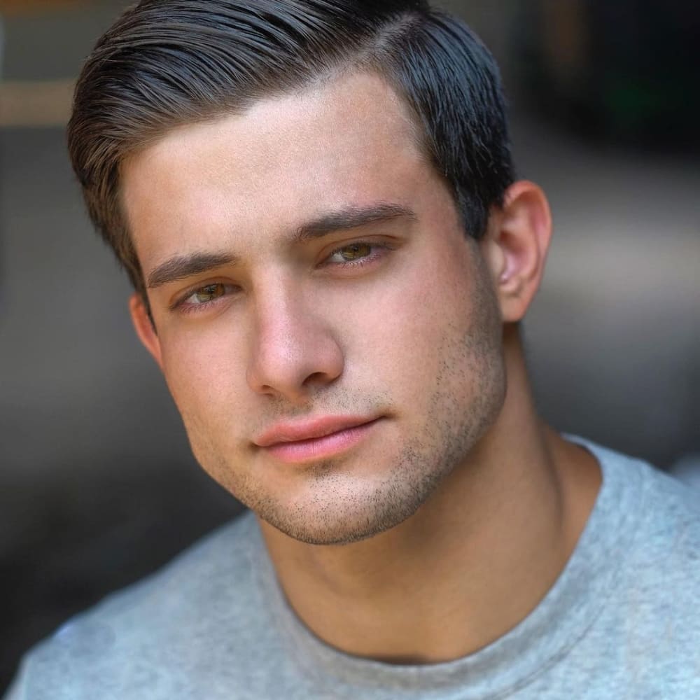 A close-up portrait of a young man with short, neatly styled dark hair and a subtle 5 o'clock shadow on his face. He is wearing a light gray shirt and has a calm, confident expression with slightly parted lips. The background is softly blurred, keeping the focus on his face.