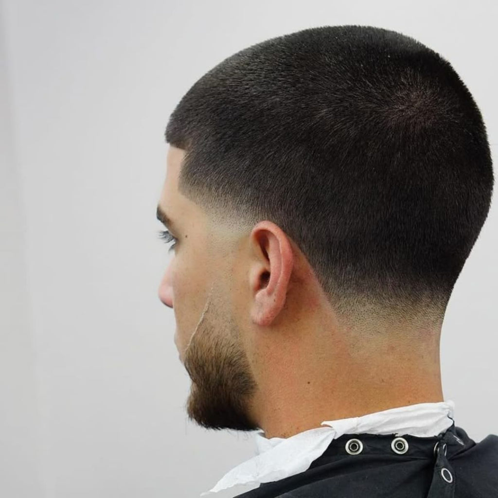 Back view of a man with a fresh buzz cut low taper haircut. The hair on top is evenly short, while the sides and back feature a smooth, gradual fade starting just above the ears and tapering down towards the neckline. The clean fade showcases a well-defined, subtle transition from hair to skin, giving a sleek and polished look.