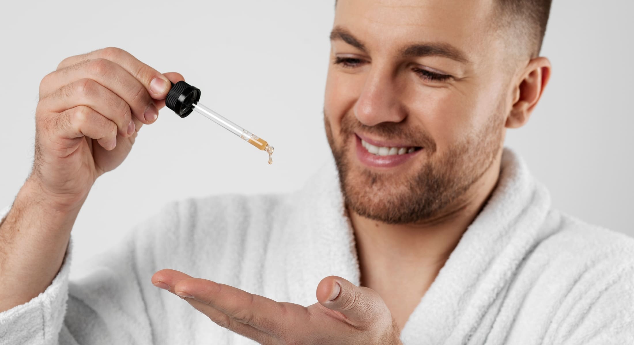 Smiling man applying beard oil with a dropper as part of his daily grooming routine, highlighting the benefits of using beard oil every day for a soft and healthy beard.