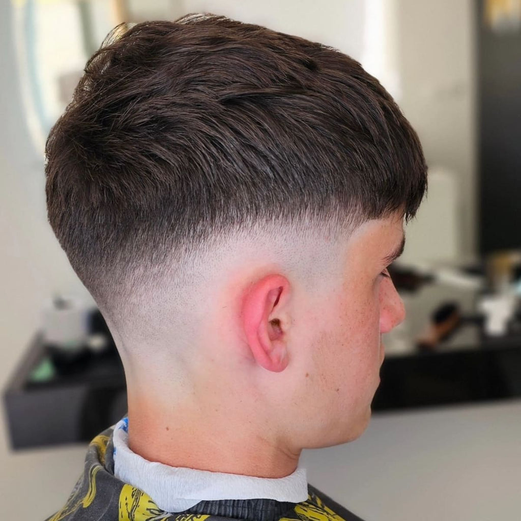 A close-up side view of a young man with a French crop mid fade haircut. The top of his hair is textured and styled forward, while the mid skin fade on the sides creates a smooth transition from the longer top to the shaved sides. The fade is clean and sharp, blending gradually from the hairline down to the nape. He is sitting in a barber’s chair, wearing a black and yellow cape, with the background blurred to emphasize the French crop mid fade hairstyle. The precise fade and textured top give the cut a stylish and modern look.