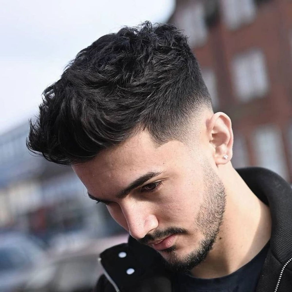 Side profile of a man with a stylish quiff haircut, featuring a textured, voluminous top and a smooth, tapered fade on the sides. The look is complemented by a short beard, adding a touch of rugged elegance to his appearance.