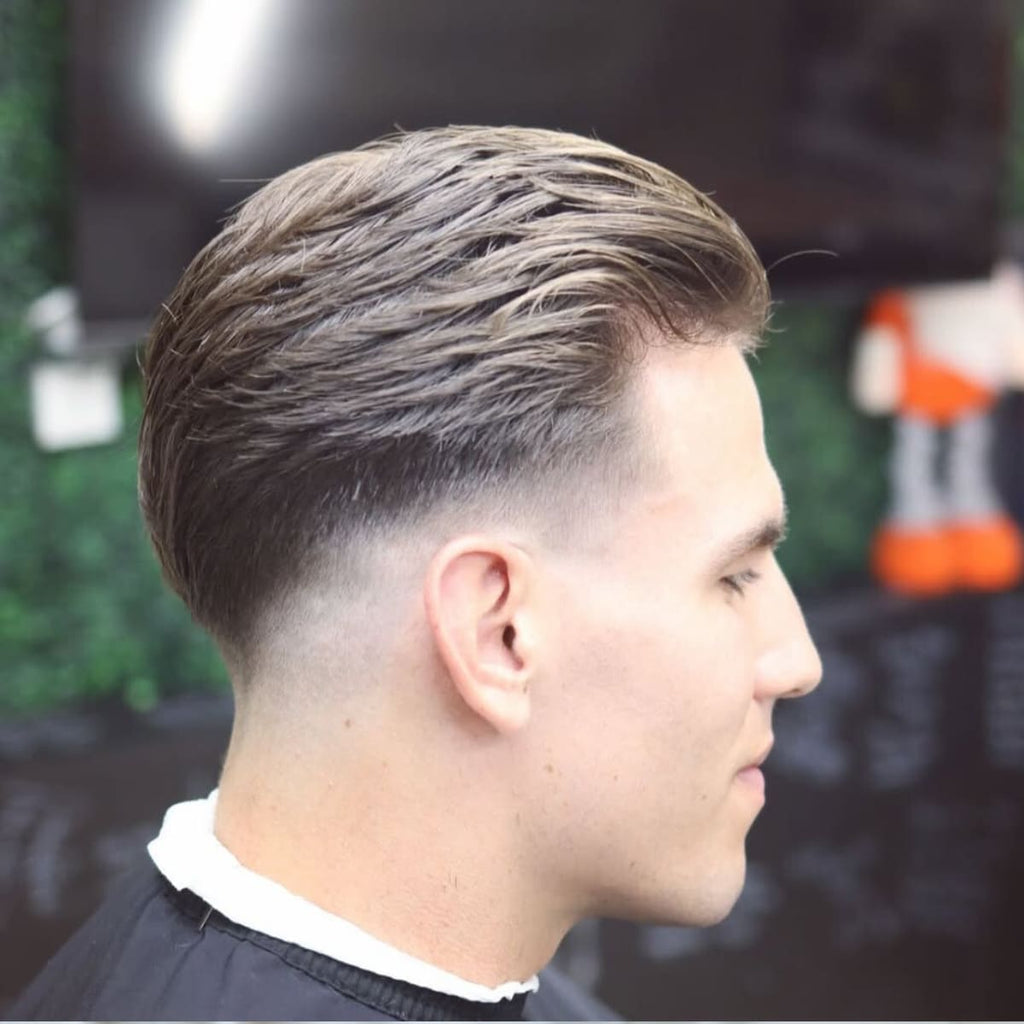 A side profile of a man in a barbershop with neatly styled hair featuring a clean taper on the sides and a textured top combed back. The sides are smooth and gradual, starting at the base of the hairline and blending seamlessly upwards. The man’s expression is calm, and the background includes a blurred green wall and subtle barbershop elements, creating a relaxed and professional atmosphere that emphasizes the haircut's sharp details.