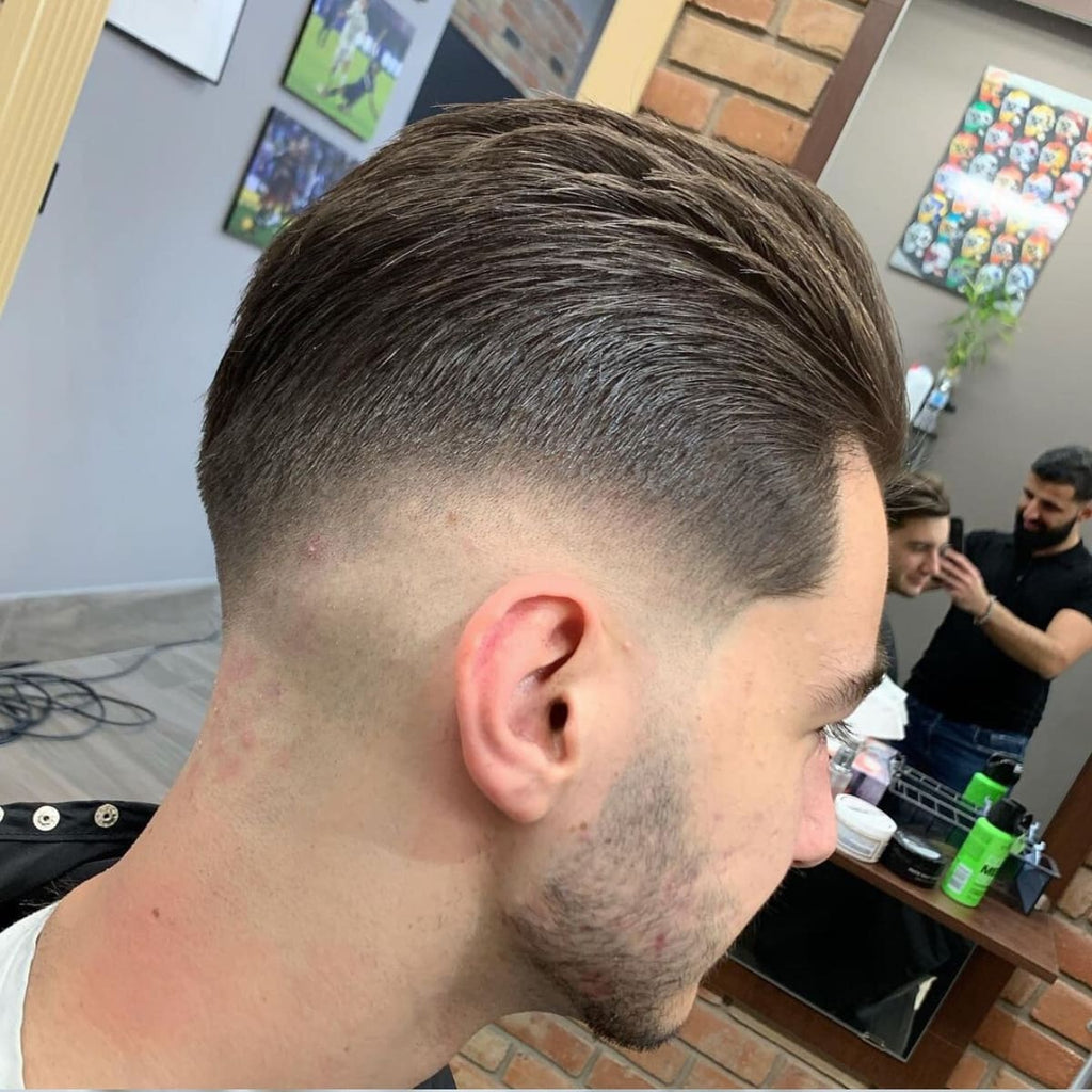 A side profile of a man in a barbershop chair with neatly styled hair, featuring a smooth blend starting low at the base of the hairline and blending gradually toward the crown. The top is combed back with a slight texture, creating a polished yet natural look. The man has light stubble on his face, complementing the haircut's sharp details. The background shows a barber working with another client, along with a mirror reflecting the haircut from the front, adding a lively and authentic barbershop atmosphere.