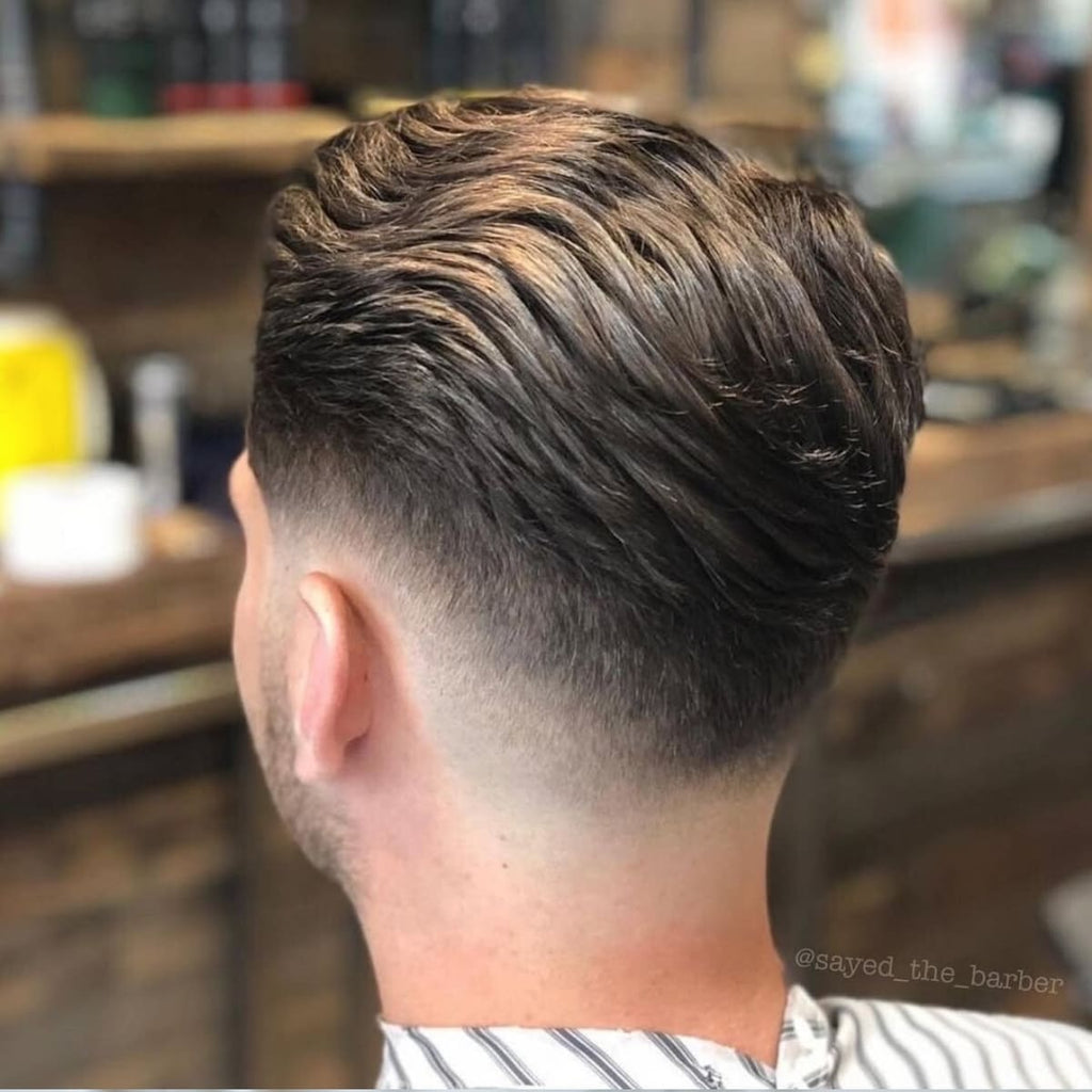 A back view of a man with a low fade slick back haircut, highlighting the smooth transition of the fade starting at the base of the neckline and gradually tapering below the ears. The top is styled with visible texture, combed back neatly for a polished and modern appearance. The background features a barbershop with wooden shelves and styling products, creating a warm and professional setting that complements the sharp details of the haircut.