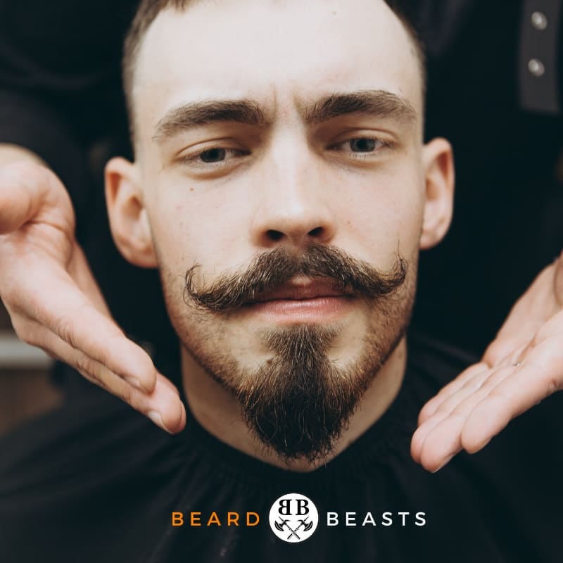 Stylish young man with one of the best Beard styles for a round face, the meticulously groomed Van Dyke beard and twirled mustache, framing his chin and lips, wearing a black turtleneck, with 'Beard Beasts' branding.