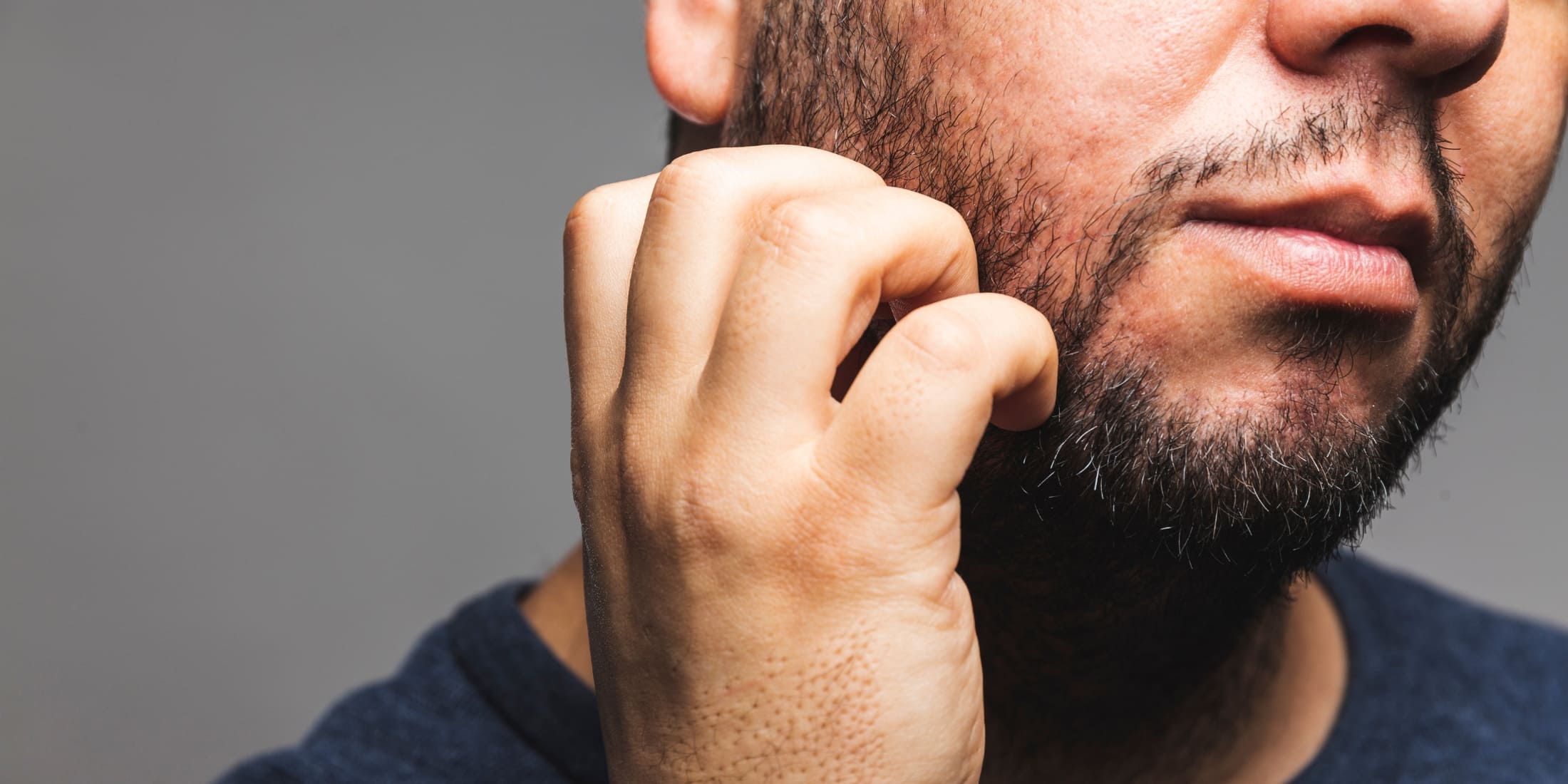 A close-up image of a man with a 2 week beard scratching his face. The man's beard is short but visibly grown in, covering his jawline and chin with some patchiness. His hand, positioned near his cheek, shows him scratching, suggesting itchiness often experienced during early beard growth stages. The background is a neutral gray, and the man is wearing a dark blue shirt. The image captures the common discomfort associated with new beard growth and the natural response to it.