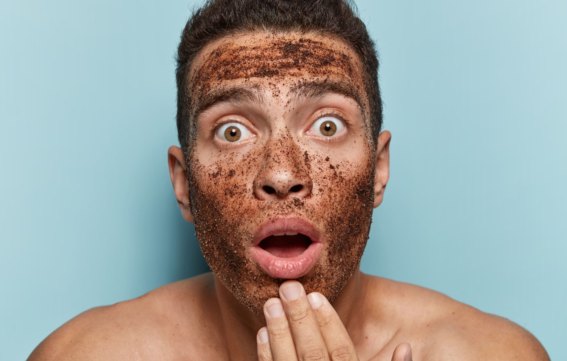 A close-up of a man with wide, surprised eyes, covered in an excessive amount of exfoliating scrub on his face. His expression is both humorous and shocked, as if he just realized he might have gone a bit overboard with the product. The scrub particles are clearly visible, emphasizing the importance of moderation in skincare tips for men.