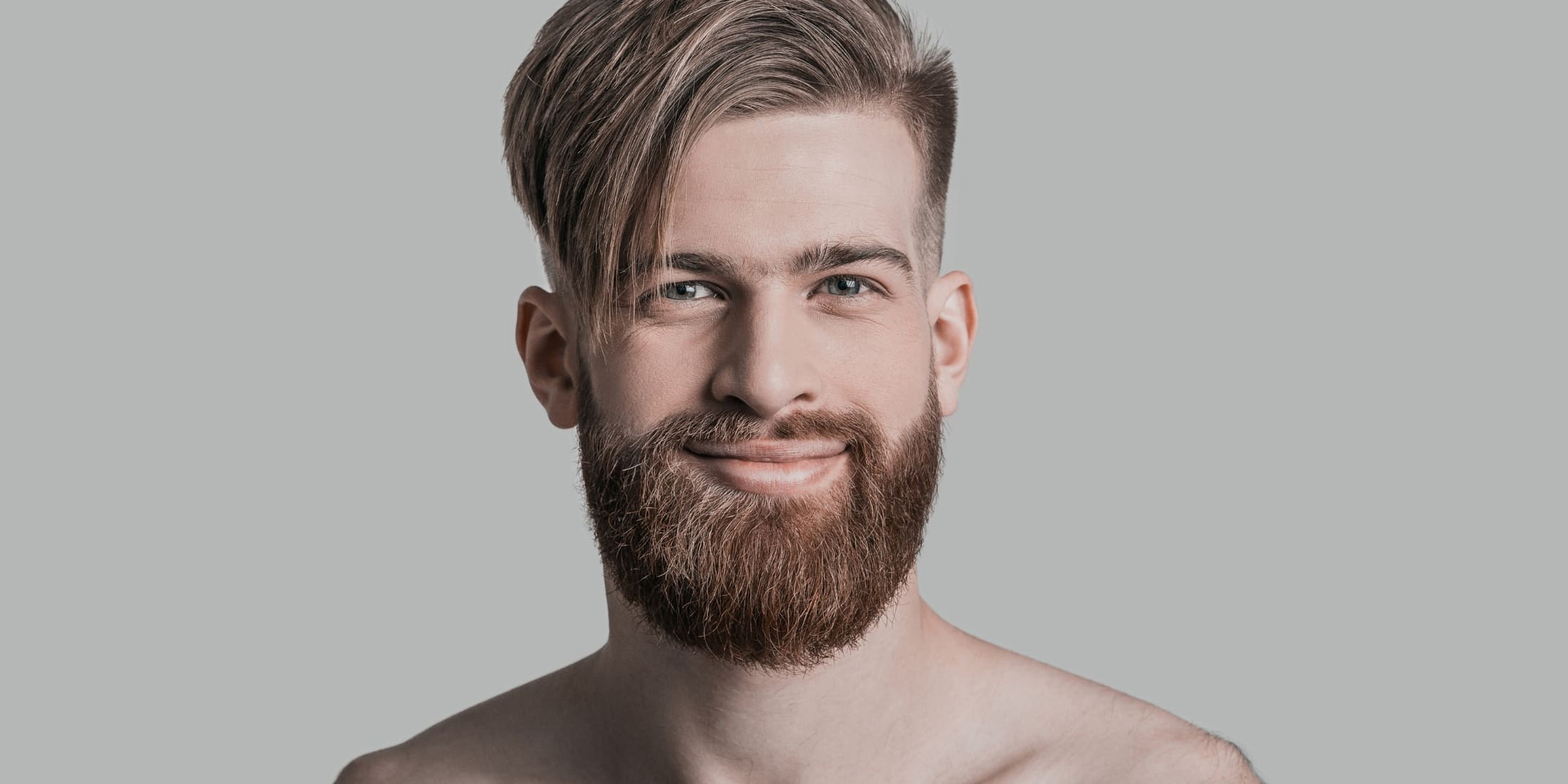 Image Alt Text: Close-up portrait of a man with a stylish, asymmetrical haircut featuring a long fringe swept to the side and a neatly groomed beard. This image highlights modern men's hair care and grooming trends, showcasing a well-maintained hairstyle and beard that reflect attention to detail and personal style. Ideal for illustrating effective hair care and grooming practices for men.