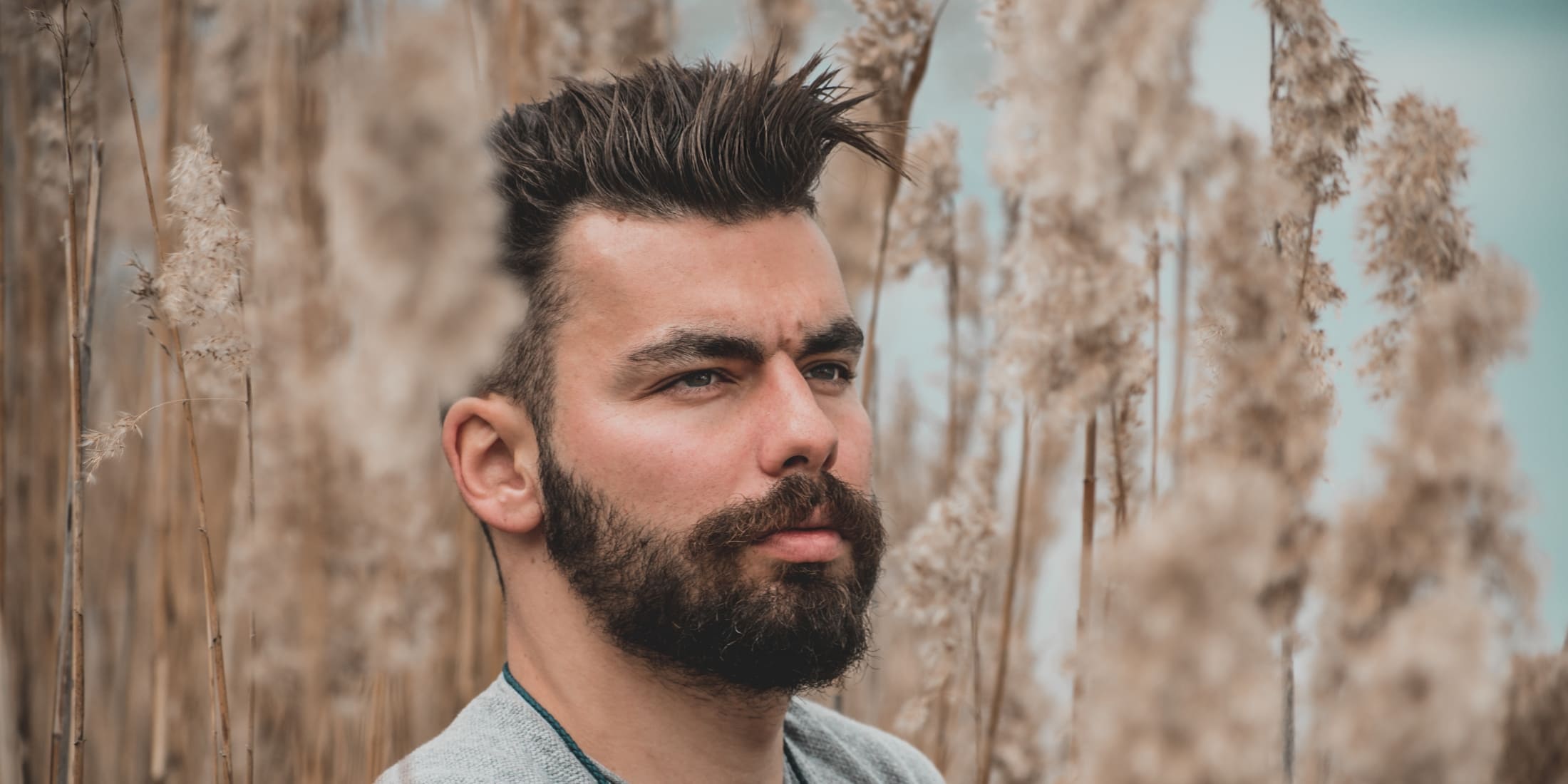 Close-up of a man with a stylish, spiked hairstyle and a full beard, standing amidst tall, dried plants. The hairstyle is voluminous and well-groomed, exemplifying modern men's hair care and grooming trends. This image is ideal for showcasing effective hair styling techniques and beard maintenance for men, highlighting attention to detail and personal style in grooming practices.