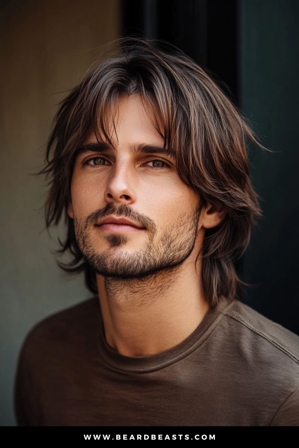 A young man sporting a trendy long shag haircut, a popular choice among layered haircuts for men. His hair flows naturally with layered, tousled strands that frame his face and add effortless texture and movement. The medium brown hair with soft highlights gives his style a sun-kissed, lived-in look, perfect for a laid-back yet stylish appearance. Complemented by light facial hair and a casual brown shirt, his look exudes a relaxed, confident vibe, enhanced by the natural lighting that highlights the texture of his layered hairstyle.