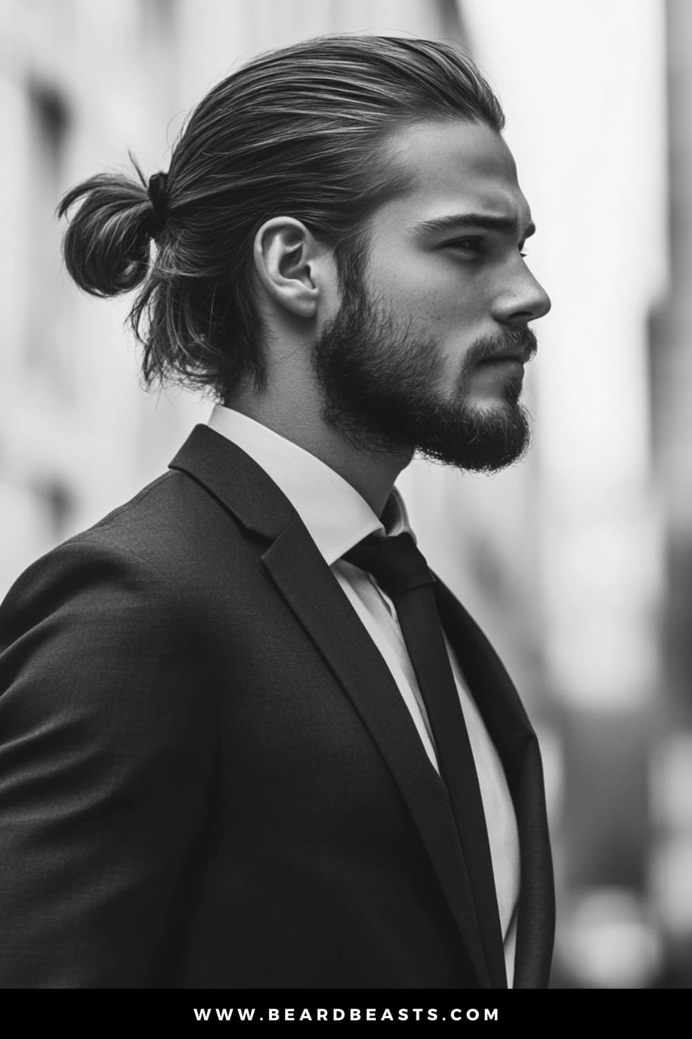 Side profile of a stylish man with a well-groomed beard and a sleek low ponytail, wearing a tailored suit and tie. One of the best men's hairstyles for long hair.