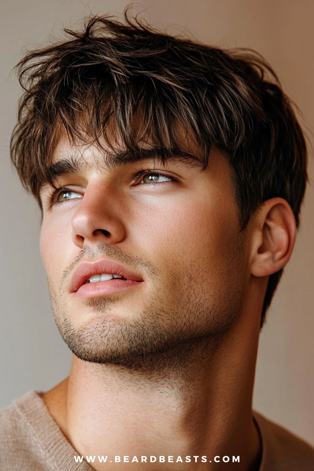 The image features a close-up of a young man with a men's shaggy hairstyle with fringe. His hair is medium-length, with soft, tousled layers that create a relaxed and effortless look. The fringe falls gently across his forehead, adding a modern, slightly edgy touch to the style. His facial hair is neatly trimmed into a light stubble, complementing the casual yet polished appearance. The overall look is trendy and youthful, perfect for those seeking a stylish yet low-maintenance haircut. The background is subtly blurred, keeping the focus on his men's shaggy haircut and the natural texture of his fringe.