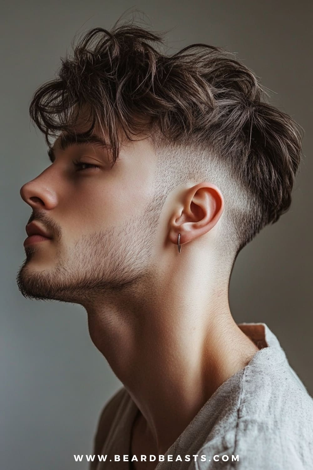 The image features a close-up side profile of a young man with a men's shaggy hairstyle with an undercut. His hair on top is styled in loose, tousled layers that add volume and texture, creating a dynamic and modern look. The sides and back of his head feature a clean, sharp undercut, providing a striking contrast to the longer, shaggy top. The undercut is precisely faded, accentuating the sharpness of the look. He has a light, well-groomed beard that complements the edgy yet stylish vibe of the hairstyle.