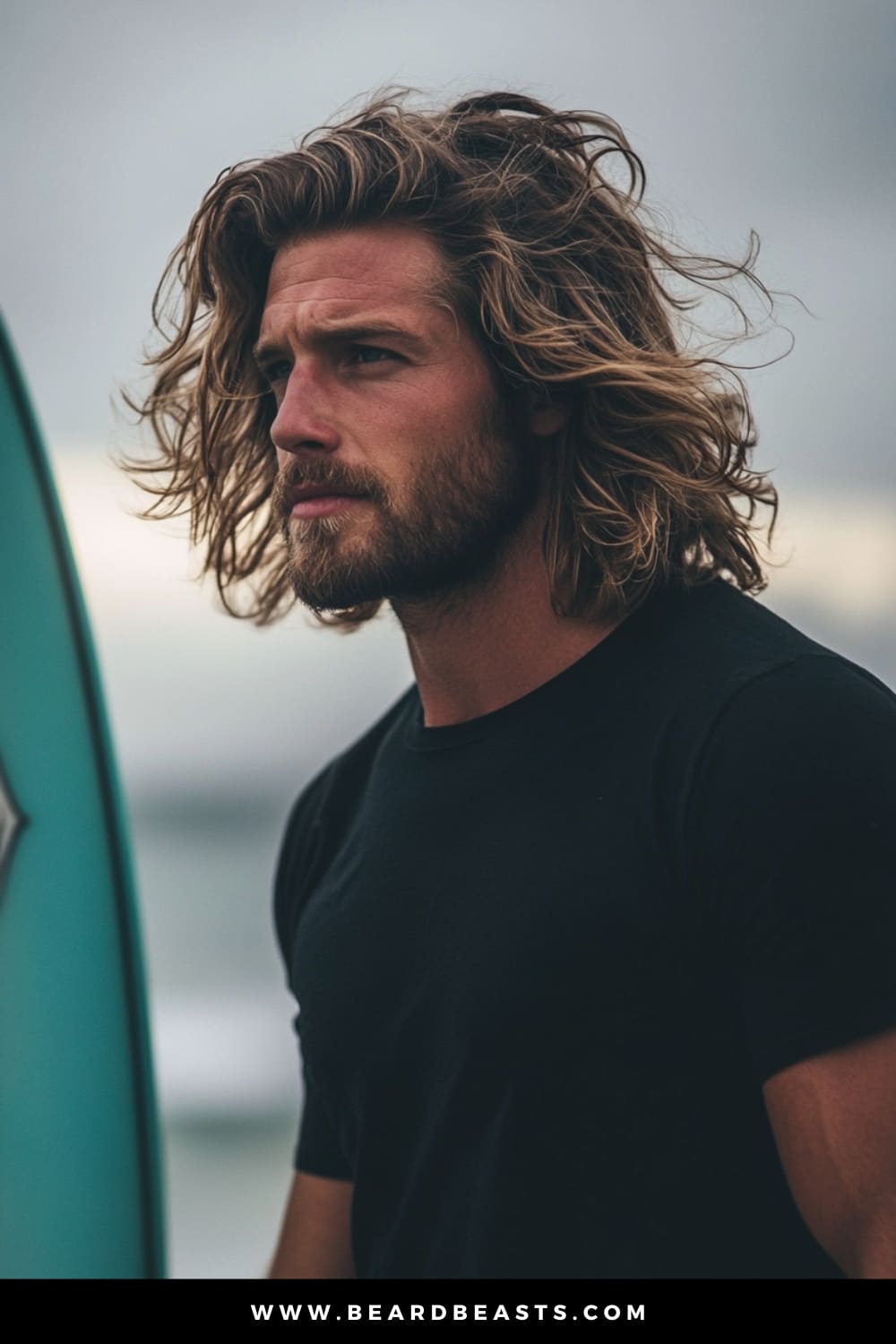 Man with textured, wavy hair and a rugged beard, standing by the beach with a surfboard, embodying the effortless, windswept look of men's hairstyles for long hair. This carefree surfer style perfectly captures a laid-back, adventurous vibe, ideal for beach lovers and outdoor enthusiasts.