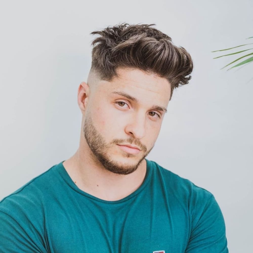 A young man with a stylish messy pompadour haircut is posing for the camera. The sides of his head are neatly faded, creating a sharp contrast with the voluminous, tousled hair on top. His hair has a natural, textured look, giving the pompadour a relaxed and casual vibe. He has a neatly trimmed beard that complements his overall appearance. Dressed in a teal t-shirt, the man exudes a laid-back yet trendy style. The background is plain white, keeping the focus on his fashionable haircut.