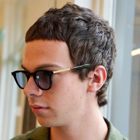 A young man is shown with a micro fringe haircut, characterized by very short, straight-across bangs that sit high on his forehead. His hair is textured and slightly wavy, styled close to the scalp with a natural, tousled look that adds a casual, effortless vibe to the overall appearance. The micro fringe creates a bold and minimalistic statement, contrasting with the slightly longer hair on the sides and back. He is wearing dark sunglasses with a thin, gold-accented frame, and his expression is relaxed and contemplative. Dressed in a green shirt, the man appears to be indoors, near a large window that lets in soft, natural light, which highlights the texture and detail of his hair. The clean lines of the micro fringe, paired with the casual waves and sunglasses, give him a modern and stylish look, ideal for someone seeking a unique and low-maintenance haircut.