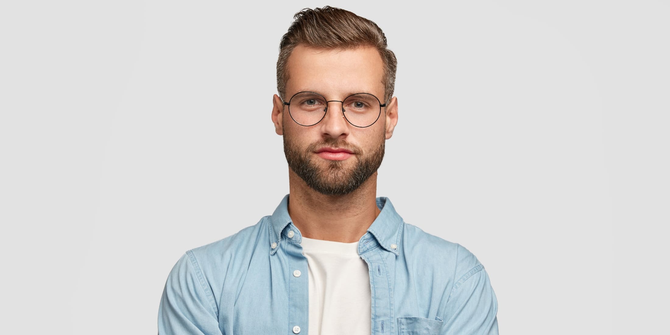 Portrait of a young man with glasses and a neatly groomed, medium-length beard, wearing a light blue shirt. The beard is in the mid-stages of growth, showcasing fullness and definition. Ideal representation of beard care during weeks 3-6, when to start using beard oil to maintain softness, manageability, and healthy skin beneath.