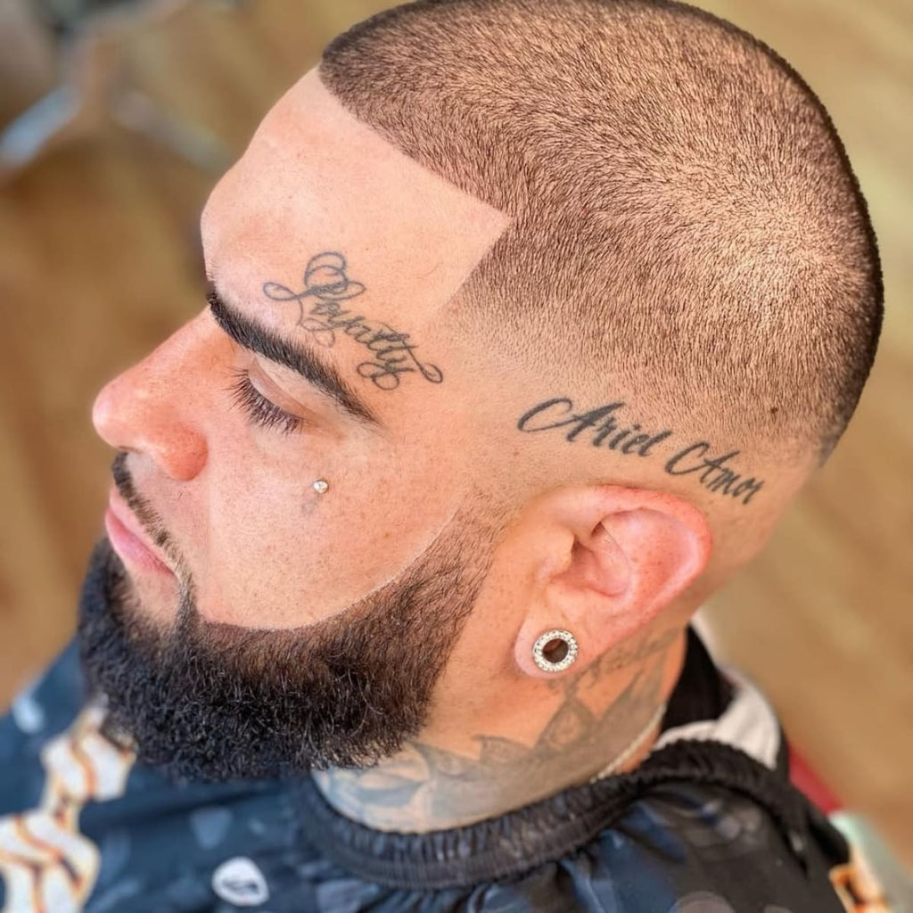 A close-up side profile of a man with a Mid Bald Fade Buzz Cut, showcasing a sharp and clean fade that blends seamlessly into a short, evenly textured top. His beard is precisely groomed, complementing the haircut, while tattoos on his forehead and neck add a unique and edgy flair.