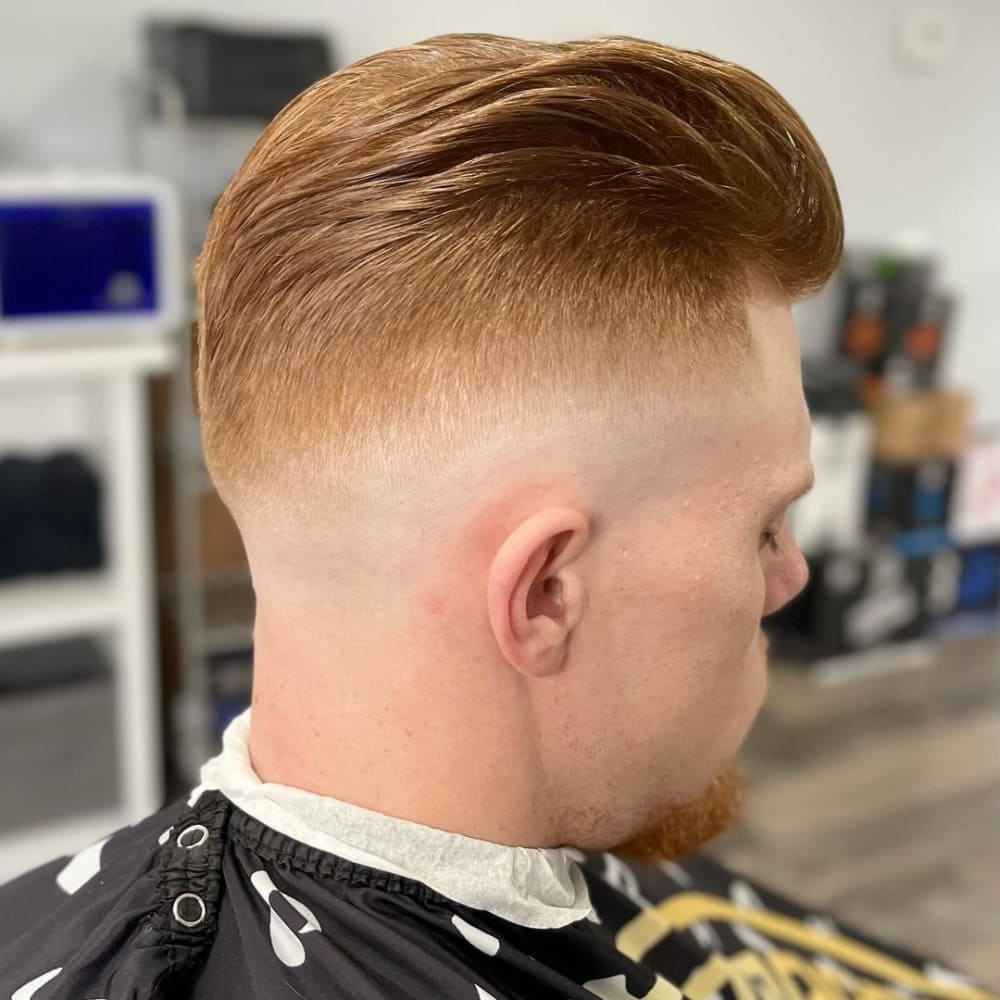 A man with a mid fade pompadour haircut sits in a barbershop chair. His hair is styled in a voluminous pompadour at the top, showcasing a smooth and sleek finish. The sides are expertly faded, transitioning seamlessly from the skin at the bottom to thicker hair towards the top. The man's hair is a striking shade of red, adding a unique flair to the classic cut. The barbershop environment is modern and clean, with organized shelves and contemporary decor, creating a professional atmosphere. The overall look is sharp and sophisticated, highlighting the precision of the fade and the fullness of the pompadour.