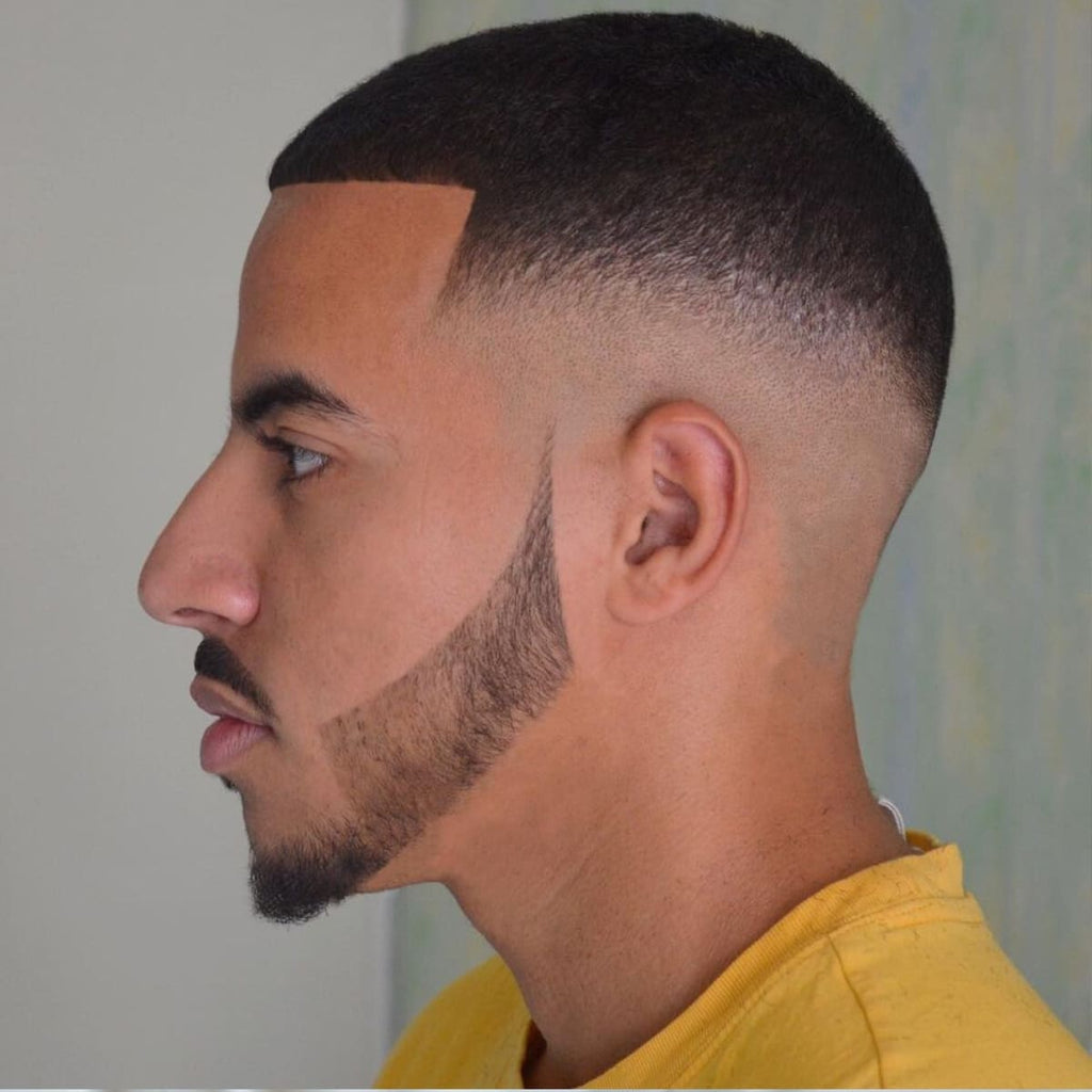 A side profile of a man with a skin fade buzz cut, featuring a smooth fade from the scalp to the short hair on top. The haircut is paired with a sharp, angular beard that blends seamlessly into the fade. The precise lines and clean edges highlight the modern and stylish look. This polished style is both bold and versatile, suitable for various occasions.