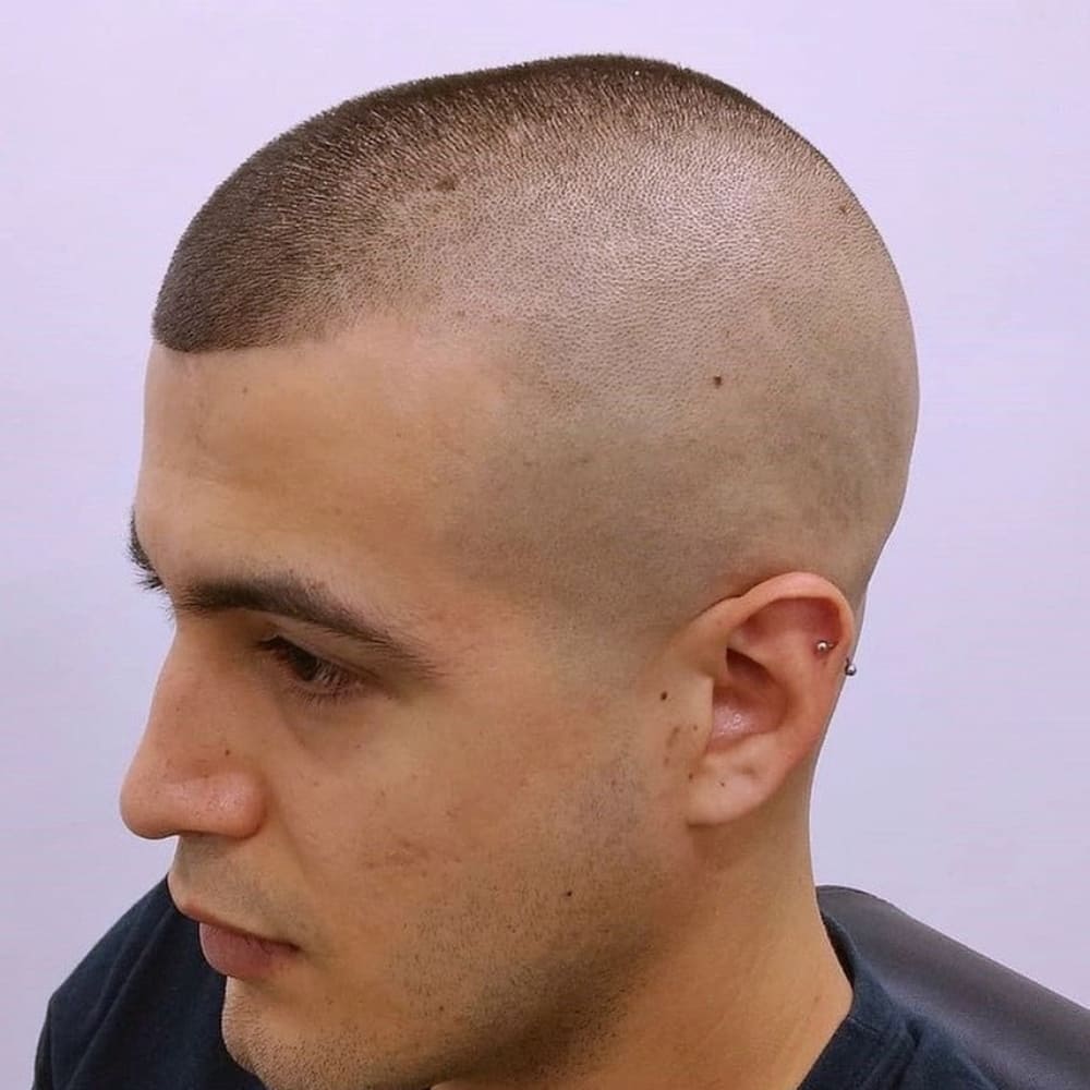 The image shows a person with a military high and tight haircut. The sides and back of the head are shaved very close to the skin, creating an almost bald appearance. The top hair is kept very short, clipped close to the scalp, and neatly trimmed to a uniform length. This haircut is characterized by its sharp, disciplined look, reflecting its military origins. The person has a clean and precise appearance, highlighting the practicality and low maintenance of the style. The overall look is rugged, no-nonsense, and ideal for those who prefer a straightforward, efficient haircut.