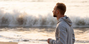 A man practicing mindfulness for men by jogging along a beach at sunrise, wearing a hoodie and listening to a mindfulness podcast through earbuds, with waves crashing in the background to reduce stress and boost mental health.