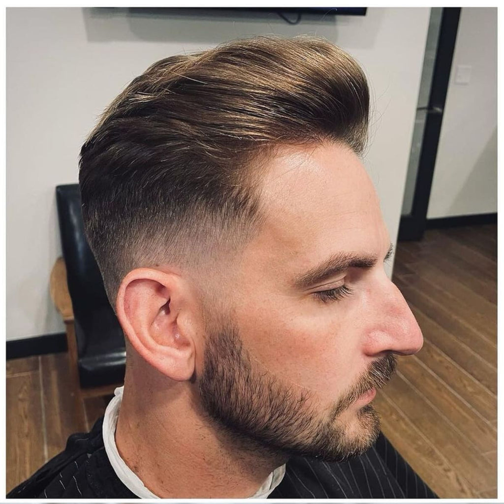 A side profile of a man with a modern low fade slick back haircut, featuring a stylishly textured top combed back with a natural, matte finish. The low fade starts at the very base of the hairline near the neckline and tapers subtly below the ears, maintaining a clean and gradual transition that enhances the haircut’s contemporary appeal. The man has a short, well-groomed beard that complements the sharp lines of the haircut. The background includes a minimalist barbershop setting with wooden floors and neutral tones, keeping the focus on the haircut's sleek and modern details.