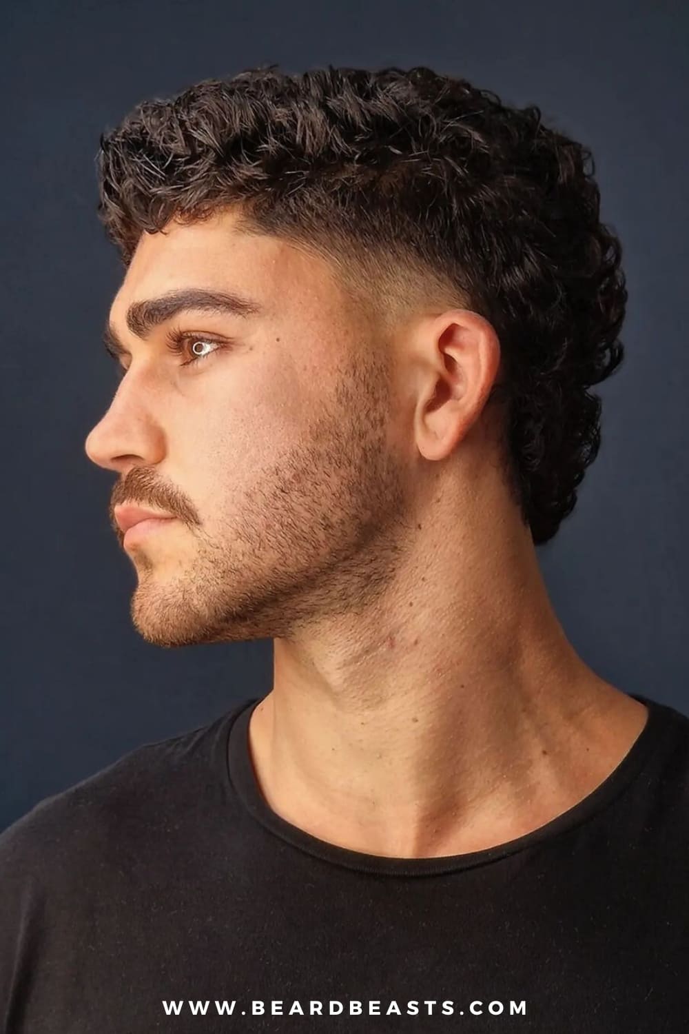 A young man with a modern mullet hairstyle is captured in profile, highlighting the sharp contrast between the close-cropped sides and the longer, textured curls at the top and back. His facial hair is neatly trimmed, complementing the overall edgy yet polished look. The dark background and simple black shirt keep the focus on the contours of his face and the defined structure of his haircut, creating a clean and striking image.