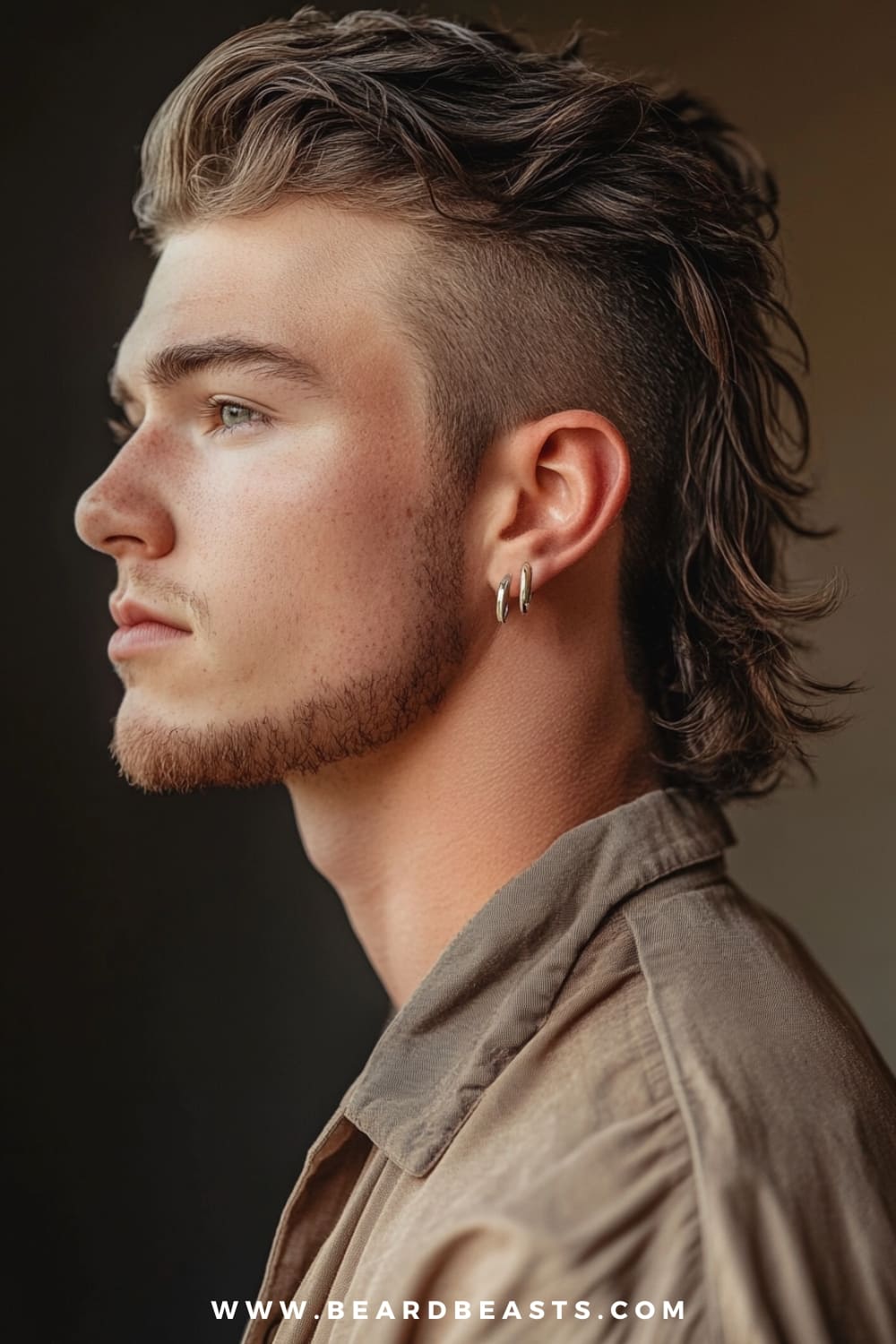 A young man is sporting a Modern Mullet, a bold and edgy take on the classic mullet, perfectly suited for those looking to make a statement with their medium length hairstyle. The style features closely cropped sides that highlight the longer, textured hair at the back, which flows naturally down the nape of the neck. The top is styled with volume and a slight wave, adding a contemporary twist to the retro-inspired cut. His look is complemented by a subtle, well-groomed beard and a pair of silver hoop earrings, enhancing the rebellious and trendy vibe of the hairstyle. The earthy tones of his shirt add to the casual yet confident aesthetic, while the softly blurred background keeps the focus on the intricate details of the mullet. This look is perfect for those who appreciate a mix of classic and modern styles with an adventurous spirit.