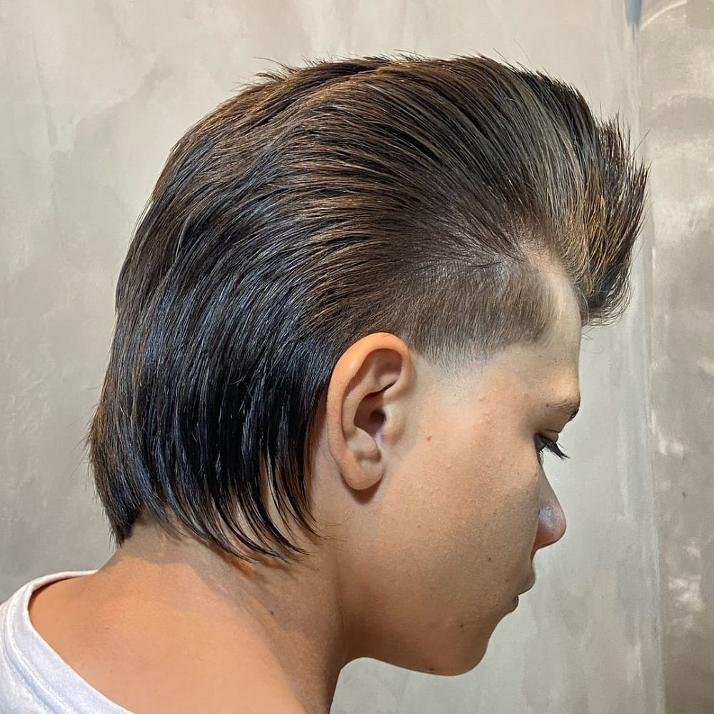 A young man is showcasing a distinctive mullet pompadour haircut. The top is styled high and slicked back, creating a voluminous pompadour effect. The sides are neatly tapered and faded, providing a clean and sharp contrast. The back features a classic mullet style, with longer hair flowing down the neck, adding a retro yet modern twist to the overall look. The man’s expression is focused, highlighting the bold and edgy nature of his unique hairstyle. The background is neutral, allowing the haircut to stand out prominently.  A network error occurred. Please check your connection and try again. If this issue persists please contact us through our help center at help.openai.com.