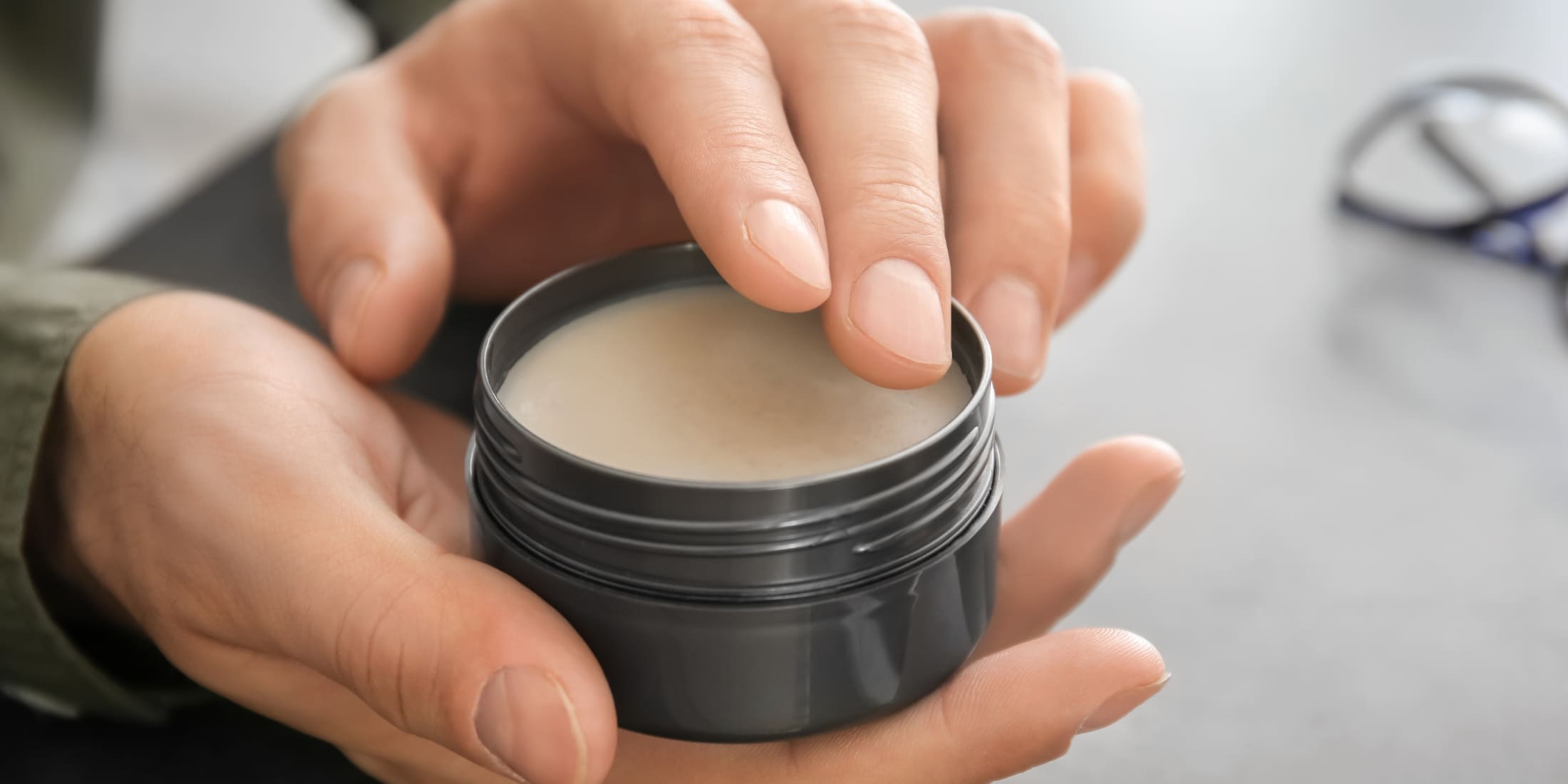 Close-up image of a person holding and applying hair pomade from a black tin, showcasing the texture and application process. Highlights the differences in texture and usability in the pomade vs wax vs clay debate.