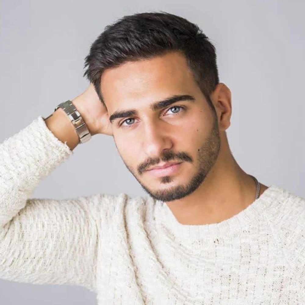 A color portrait of a young man with a neatly groomed royale beard. His beard features a thin chin strip connected to a well-defined mustache, creating a sophisticated and clean look. The beard is short and precisely trimmed, enhancing his sharp jawline and strong chin. The man has short, dark hair, styled neatly and slightly tousled. He is wearing a light, textured sweater, which contrasts nicely with his tanned skin. His expression is relaxed and confident, with one hand resting behind his head, showcasing a silver wristwatch. The background is a plain, light gray, ensuring that the focus remains on his facial features and beard style. The lighting is even and soft, highlighting the texture and grooming of his beard, giving the portrait a fresh and polished appearance.