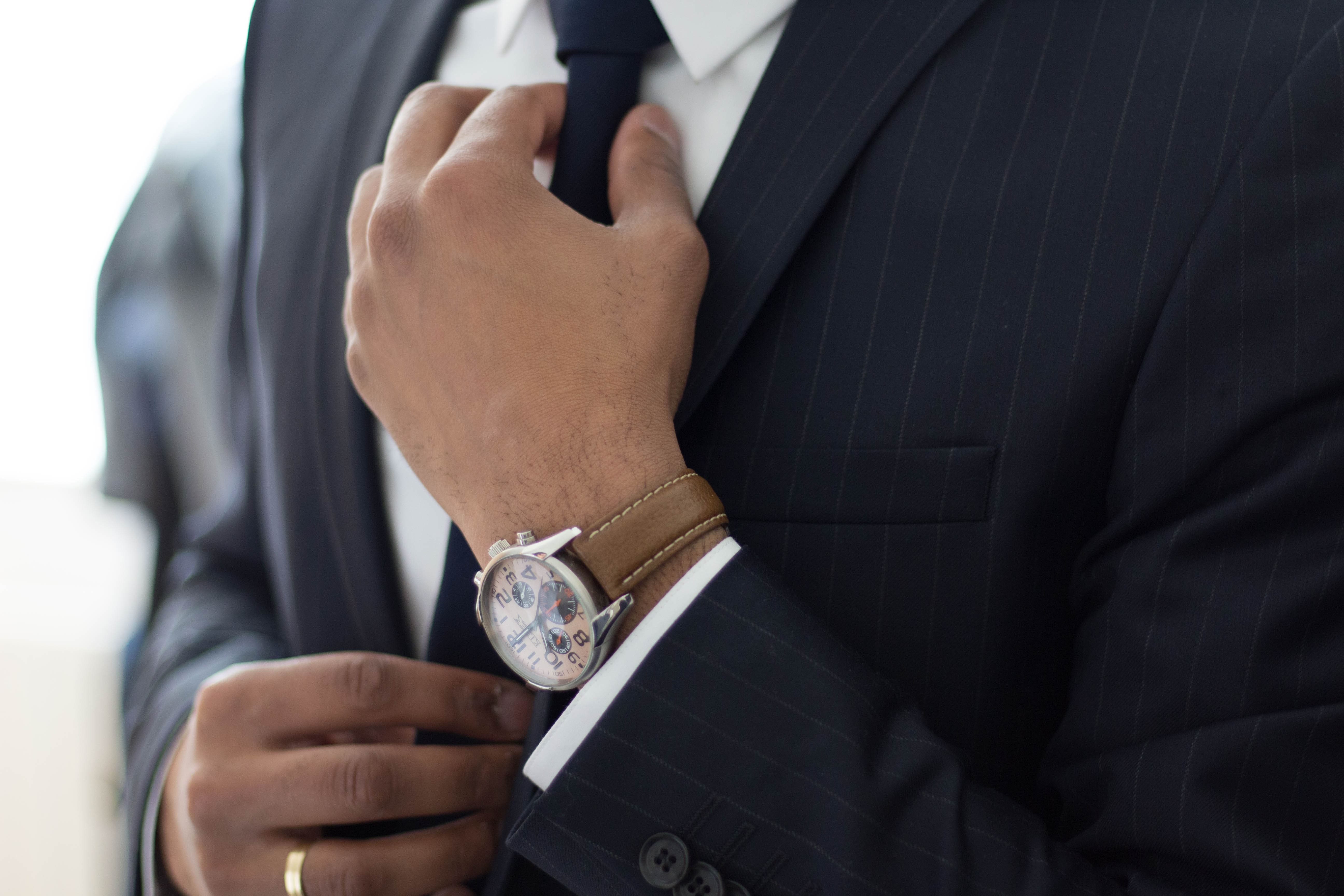 Close-up of a man's torso in a tailored navy blue pinstripe suit adjusting his black tie. His left hand, adorned with a gold wedding band, also displays an elegant wristwatch with a brown leather band. The focus on the watch and his well-groomed appearance evoke a sense of sophistication, possibly highlighting the watch as an exemplary gift for a husband.