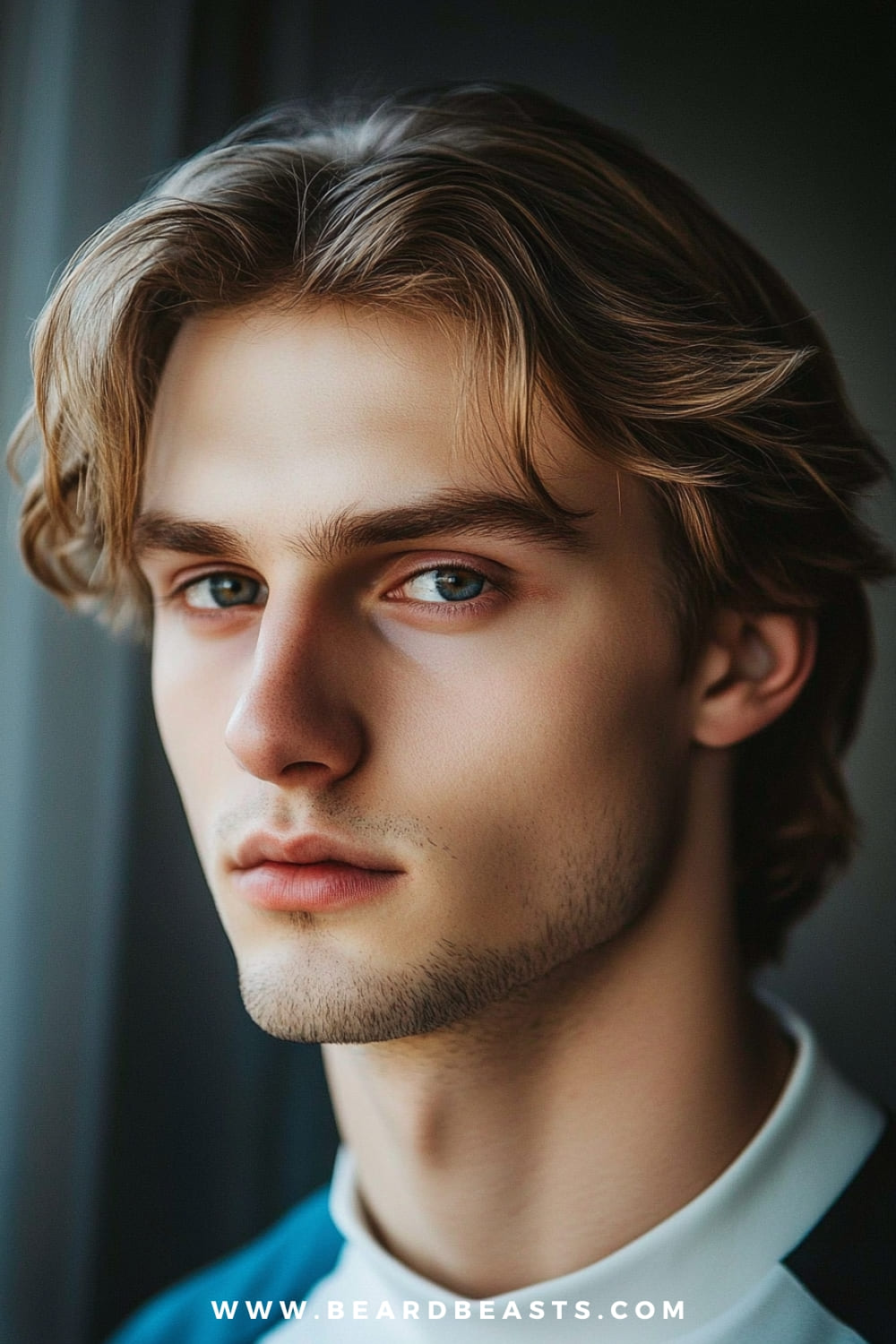 The image features a close-up of a young man with a shaggy hairstyle with a middle part. His hair is medium-length, with soft, natural waves that fall gracefully on either side of his face, creating a balanced and symmetrical look. The middle part adds a classic and refined touch, while the shaggy texture maintains a casual, relaxed vibe. His facial hair is minimal, with a light stubble that complements his youthful and fresh appearance. The lighting is soft, highlighting the natural shine and texture of his hair, while the neutral background ensures the focus remains on his hairstyle and expressive eyes. The overall look is stylish, modern, and effortlessly sophisticated.