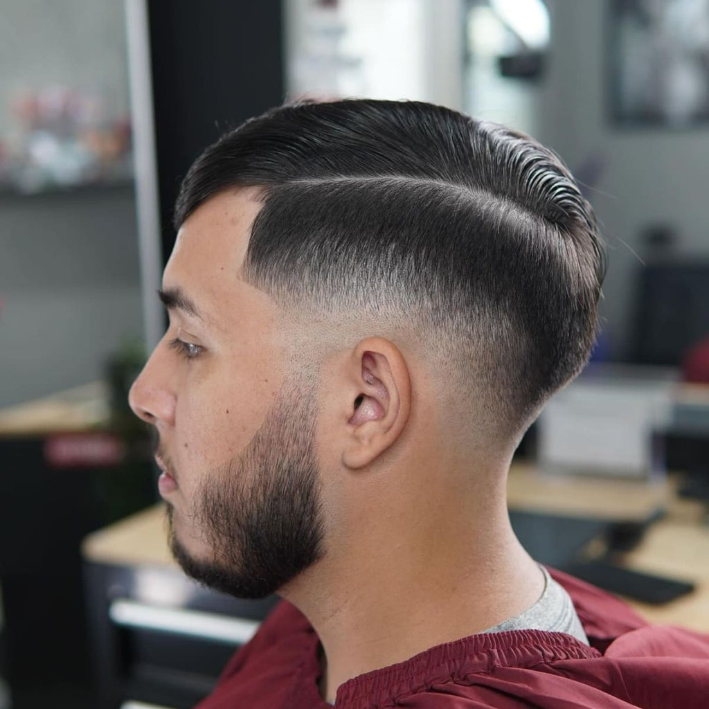 A side profile of a man with a clean, sharp comb over low fade hairstyle. The hair is neatly parted and combed over to one side, with a smooth low fade blending into the skin around the ears and neckline. The beard is well-groomed, adding to the overall polished and stylish look.