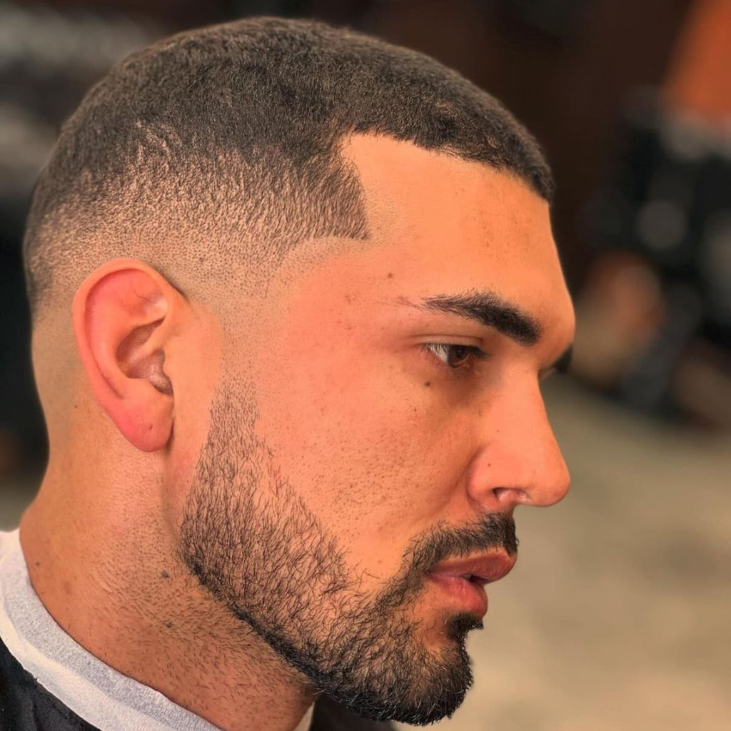 Close-up of a man with a mid fade buzz cut, featuring a sharp, gradual fade that enhances his facial structure. The haircut transitions smoothly from a short buzz on the sides to a slightly longer length on top. His defined beard line complements the mid fade, creating a clean and stylish look. Ideal hairstyle choice for men seeking a modern, low-maintenance cut with sharp detailing.