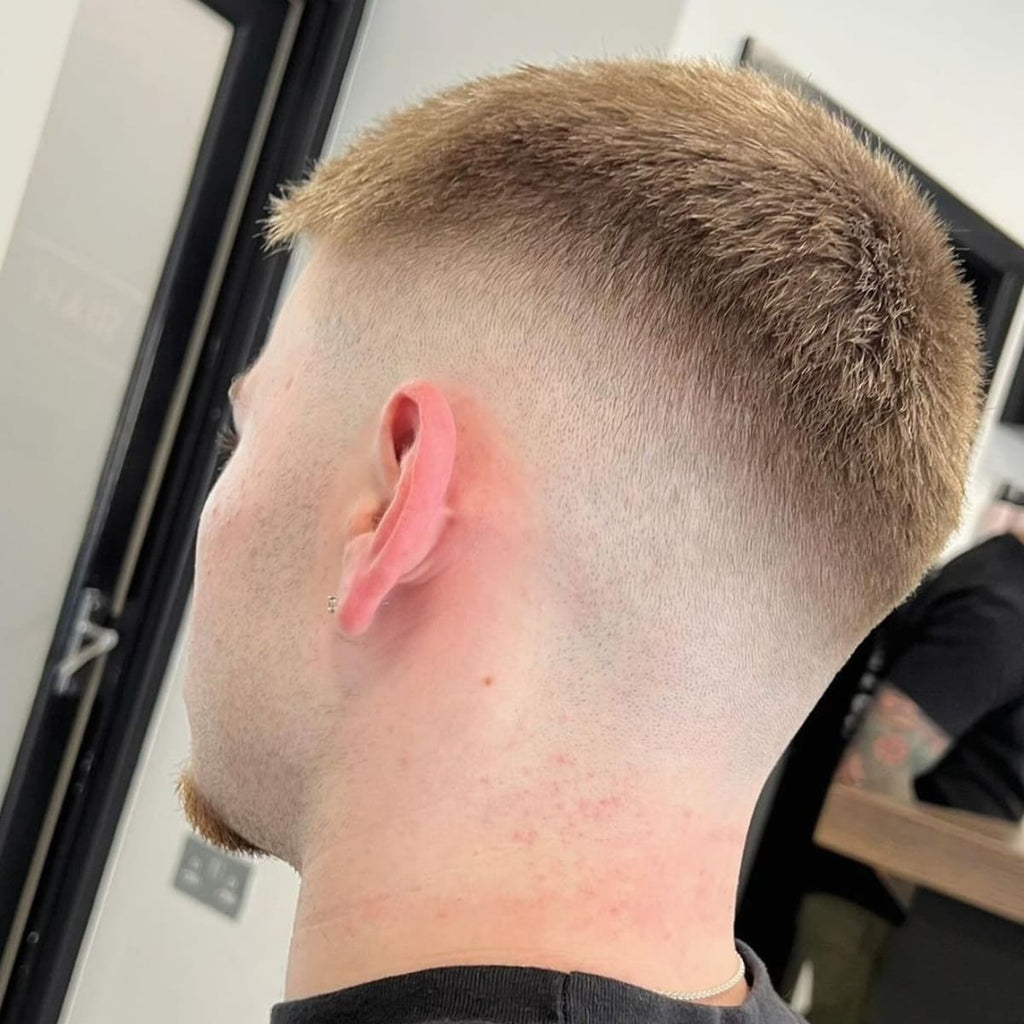 A close-up image of a man with a crew cut paired with a drop fade. The fade curves down behind the ears, creating a clean and smooth gradient from the longer hair on top to the skin fade near the neckline. The top is neatly trimmed, showcasing a short and textured finish, while the drop fade offers a sharp, modern look. The hairstyle is fresh and precise, perfect for a low-maintenance yet stylish appearance.