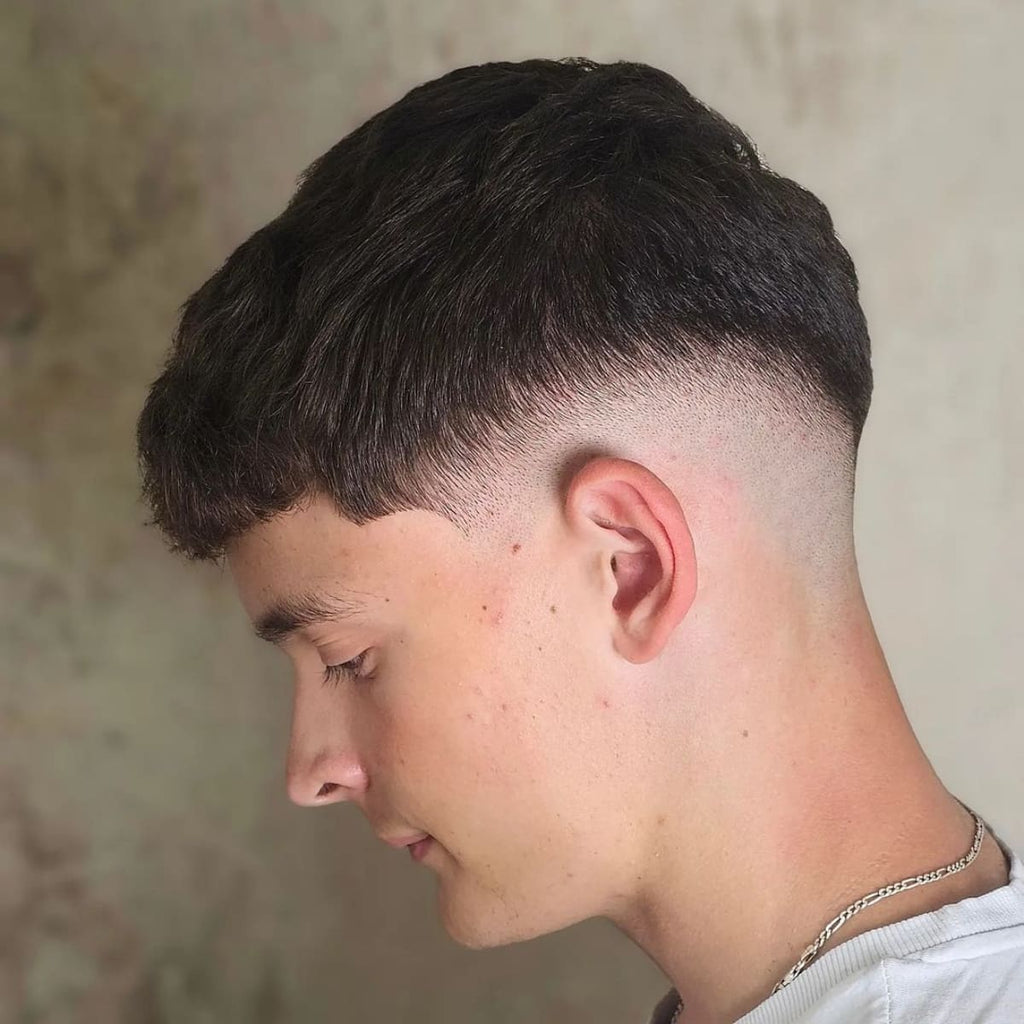 A side view of a young man sporting a French crop mid fade haircut. The top is styled with short, textured hair that blends seamlessly into a mid skin fade, starting around the temple and gradually tapering down to the nape. The smooth transition between the textured top and the skin fade gives the haircut a sharp, modern look. The man is wearing a white t-shirt and a silver chain, with a soft, neutral background that enhances the focus on his French crop mid fade hairstyle. The clean, precise fade adds a polished and stylish finish to the overall look.