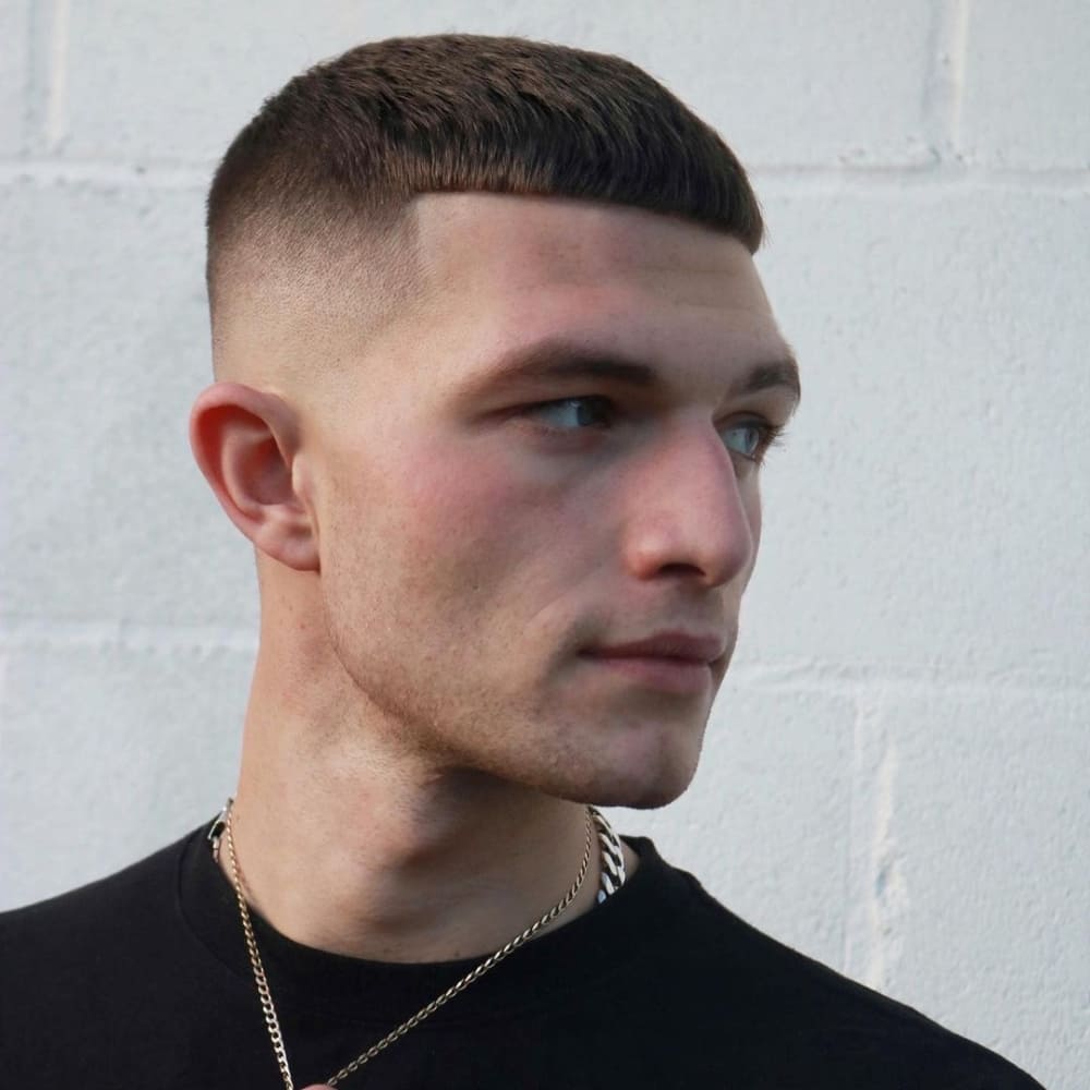 Man with a short Caesar haircut featuring a neatly trimmed, straight fringe and a high fade on the sides and back. He is wearing a black shirt and a gold chain necklace, creating a clean, sharp, and modern look against a white wall background.