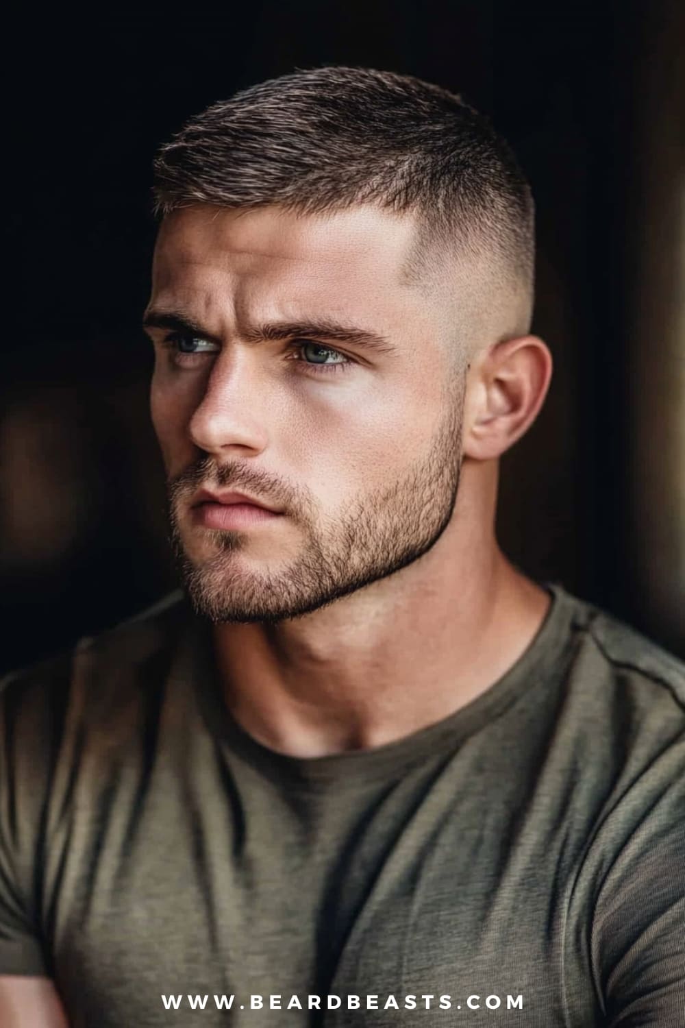 A close-up image of a man with a Short Crew Cut hairstyle, featuring neatly trimmed hair on the sides and back with a slightly longer top that is meticulously styled for a clean and structured look. The haircut exudes a classic, masculine appeal, making it one of the most popular military haircuts for men. The man also has a well-groomed, short beard that enhances his strong facial features, particularly his jawline. He is wearing an olive-green t-shirt that complements the military-inspired aesthetic of his haircut. The background is dark and softly blurred, ensuring that the focus remains on the Short Crew Cut, highlighting its simplicity, versatility, and timeless style.