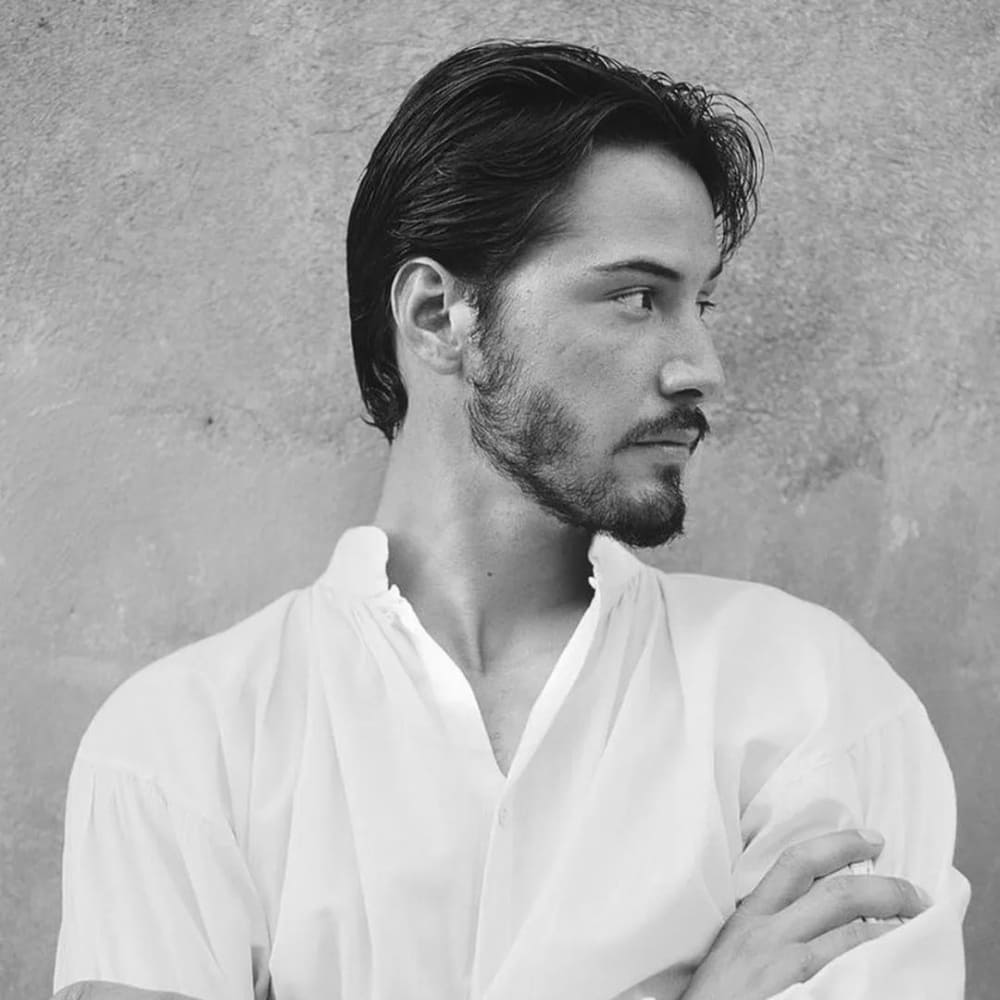 A black-and-white profile portrait of a young man with a short patchy beard. His beard is sparse in some areas, giving it a natural, uneven appearance that adds a unique character to his look. The beard is short and neatly groomed, highlighting his defined jawline and cheekbones. He has medium-length, dark hair that is slicked back, enhancing his sharp and refined appearance. The man is wearing a light, loose-fitting shirt with an open collar, contributing to a relaxed and slightly bohemian style. His expression is serious and contemplative, with his arms crossed in front of him, suggesting a thoughtful demeanor. The background is a textured wall, adding depth and interest to the portrait while keeping the focus on his facial features and beard. The lighting is soft, casting gentle shadows that emphasize the texture and natural growth pattern of his beard.