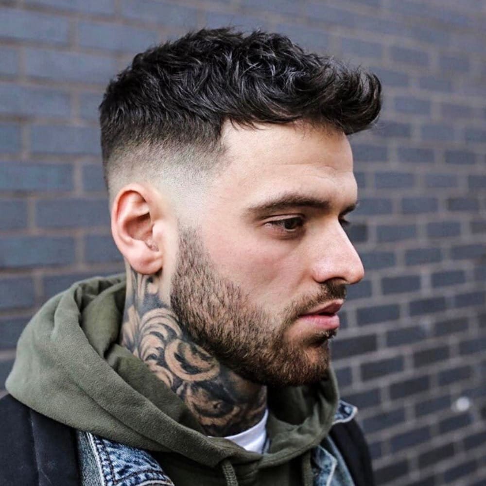 A man with a neatly groomed beard and prominent neck tattoos sports a stylish haircut. The hairstyle is a short quiff paired with a mid fade, creating a balanced and modern look. The hair on top is textured and slightly tousled, while the mid fade gradually tapers down to the skin around the ears and back. He is wearing a green hoodie and a denim jacket, standing against a dark brick wall. The overall appearance is sharp and trendy, emphasizing both the haircut and his well-maintained facial hair.