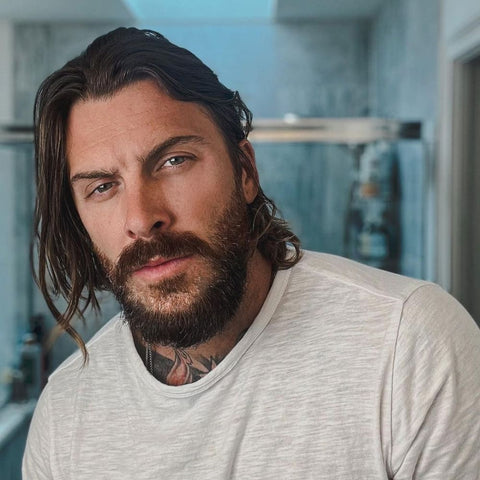 A color portrait of a man with a well-maintained short shaggy beard. His beard is full and slightly unkempt, giving him a rugged yet stylish appearance. The beard is dense and covers his face evenly, blending naturally with his long, wavy hair that falls around his shoulders. He has a piercing gaze with light-colored eyes and a confident expression. The man is wearing a plain white t-shirt, which contrasts with his dark hair and beard. Visible tattoos peek out from under his shirt at the neckline, adding to his edgy look. The background is an indoor setting, possibly a bathroom, with soft, natural light illuminating his face and enhancing the texture of his beard and hair. The overall impression is of a modern, effortlessly cool style.