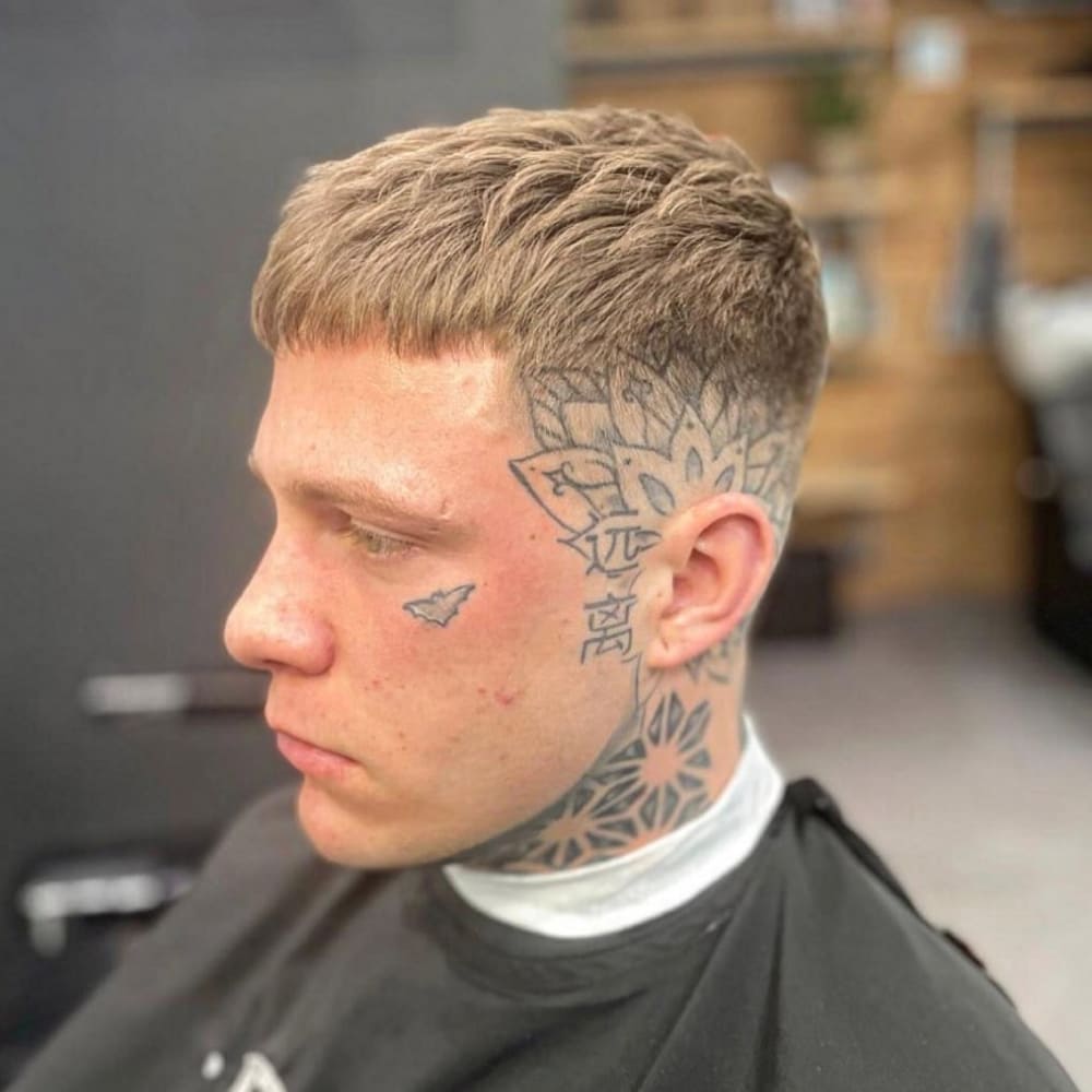 A man with a short textured crop haircut is featured in the image. His hair is cut short and styled with textured layers, creating a neat yet dynamic look. The haircut is complemented by a clean fade on the sides, which emphasizes the textured top. He has distinctive facial and neck tattoos, adding a unique and edgy element to his overall appearance. The setting appears to be a barbershop, highlighting the freshly done and well-groomed hairstyle. The man's serious expression showcases the bold and stylish nature of the short textured crop.