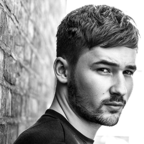 A young man is depicted with a side-swept fringe haircut, where his dark, slightly textured hair is styled neatly to the side. The fringe is longer at the front, softly sweeping across his forehead, while the sides are tapered to create a clean, sharp contrast. His hair is styled to enhance the natural texture, giving it a slightly tousled yet controlled look. The man has a well-groomed beard that adds to his rugged and masculine appearance. He is dressed in a simple, dark shirt and is leaning against a brick wall, which adds an urban and edgy element to the image. His expression is intense and focused, with a hint of seriousness in his gaze, complementing the overall style. The black-and-white effect of the photograph emphasizes the texture and contrast in his hair and beard, giving the image a classic and timeless feel.