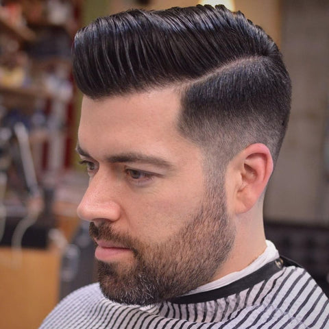 A man with a well-groomed beard and a meticulously styled side part haircut paired with a mid fade. The hair on top is neatly combed to the side, creating a polished and classic look. The mid fade begins at the middle of the head, blending smoothly into shorter hair at the sides and back. The side part is sharply defined, adding to the clean and sophisticated appearance. The man is wearing a striped barber cape, indicating he is in a barbershop. The background is slightly blurred, focusing on the precise details of the haircut. The overall style is professional and stylish, suitable for both casual and formal settings.