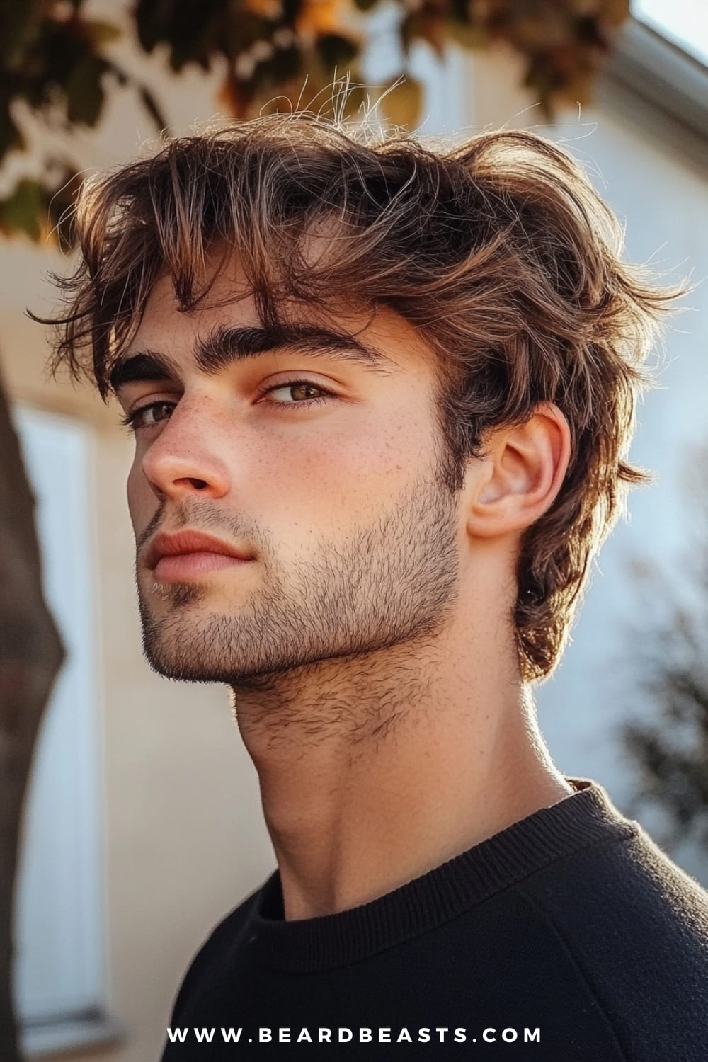 The image features a close-up side profile of a young man with a Skater Shaggy Hairstyle. His hair is medium-length, with loose, tousled layers that add volume and movement, perfectly capturing the laid-back, effortlessly cool vibe associated with skater culture. The strands of hair around his forehead and temples are slightly windswept, giving the impression of natural, carefree styling. His facial hair is a light, neatly trimmed stubble that complements the relaxed yet stylish look. The outdoor setting provides natural lighting, which highlights the texture and subtle highlights in his hair. The background, with blurred trees and a soft focus on a building, further enhances the casual, everyday feel of the image. The overall look is youthful, vibrant, and effortlessly trendy, ideal for someone with an adventurous, active lifestyle.