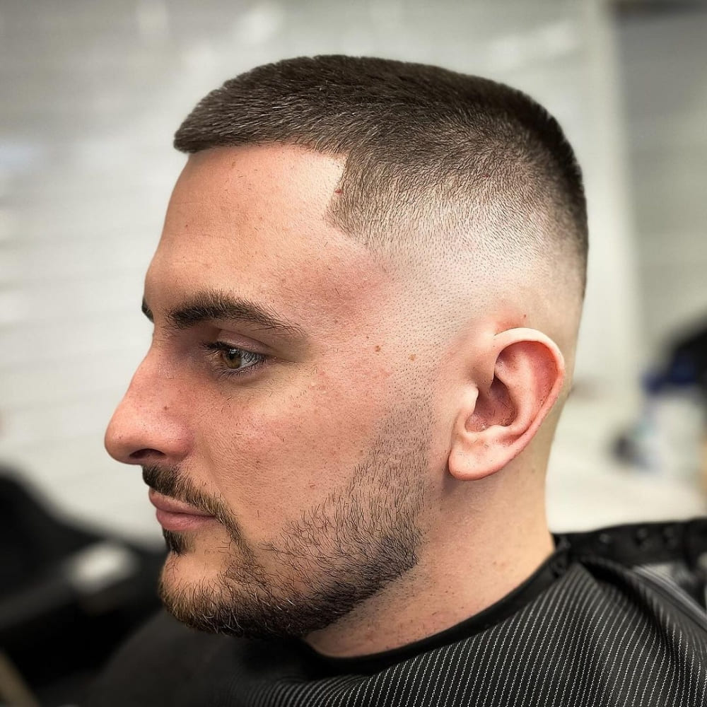 A side profile of a man with a skin fade buzz cut. The haircut features very short, neatly trimmed hair on the top that gradually fades into the skin on the sides and back, creating a clean and smooth transition. The man also has a well-groomed beard that complements the sharp and polished look of the haircut. The overall style is modern, sleek, and emphasizes the crisp edges around the hairline and beard.